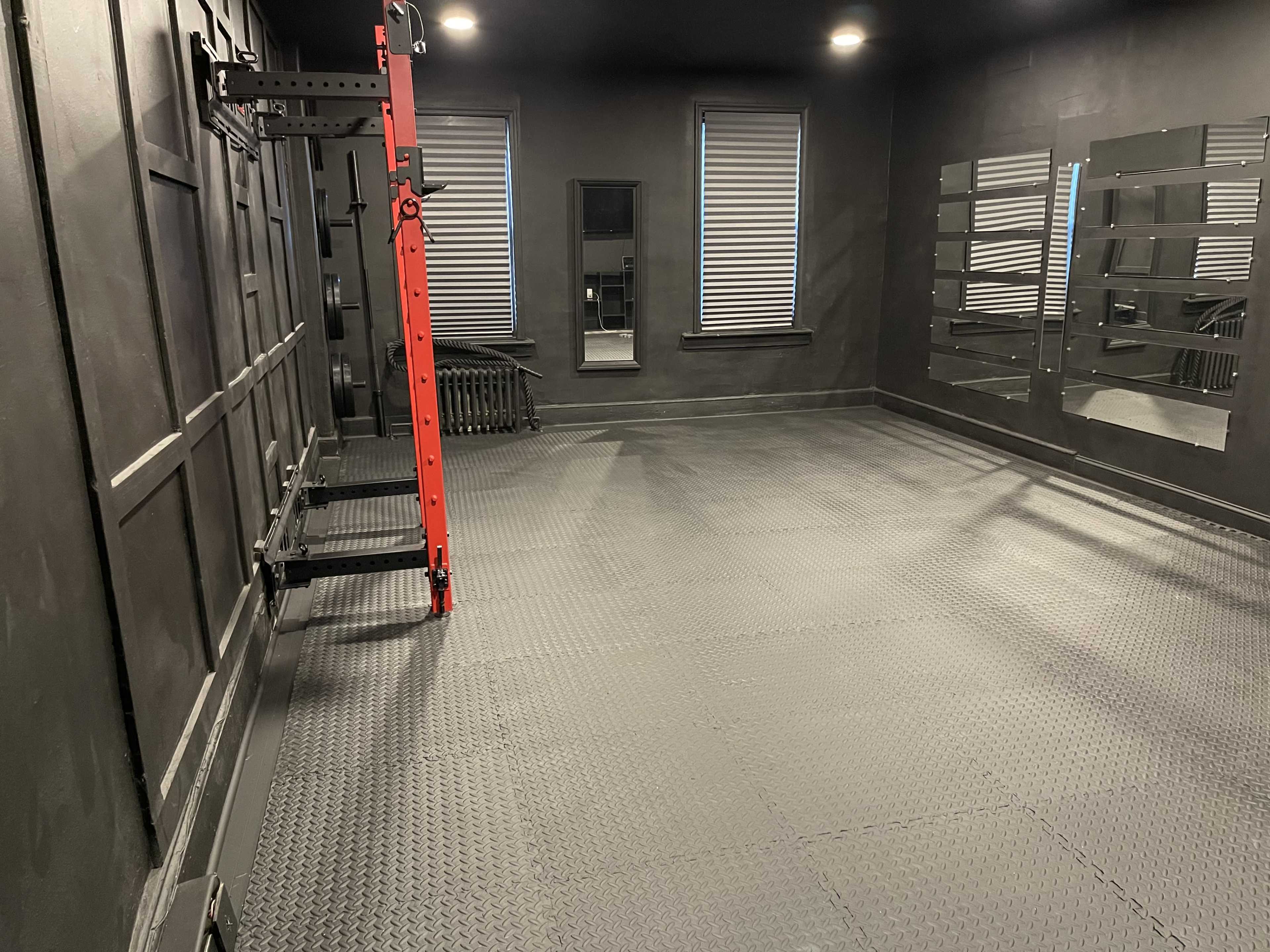 An empty, dark-colored gym interior with a red workout rack on one wall and large mirrors on the opposite wall.