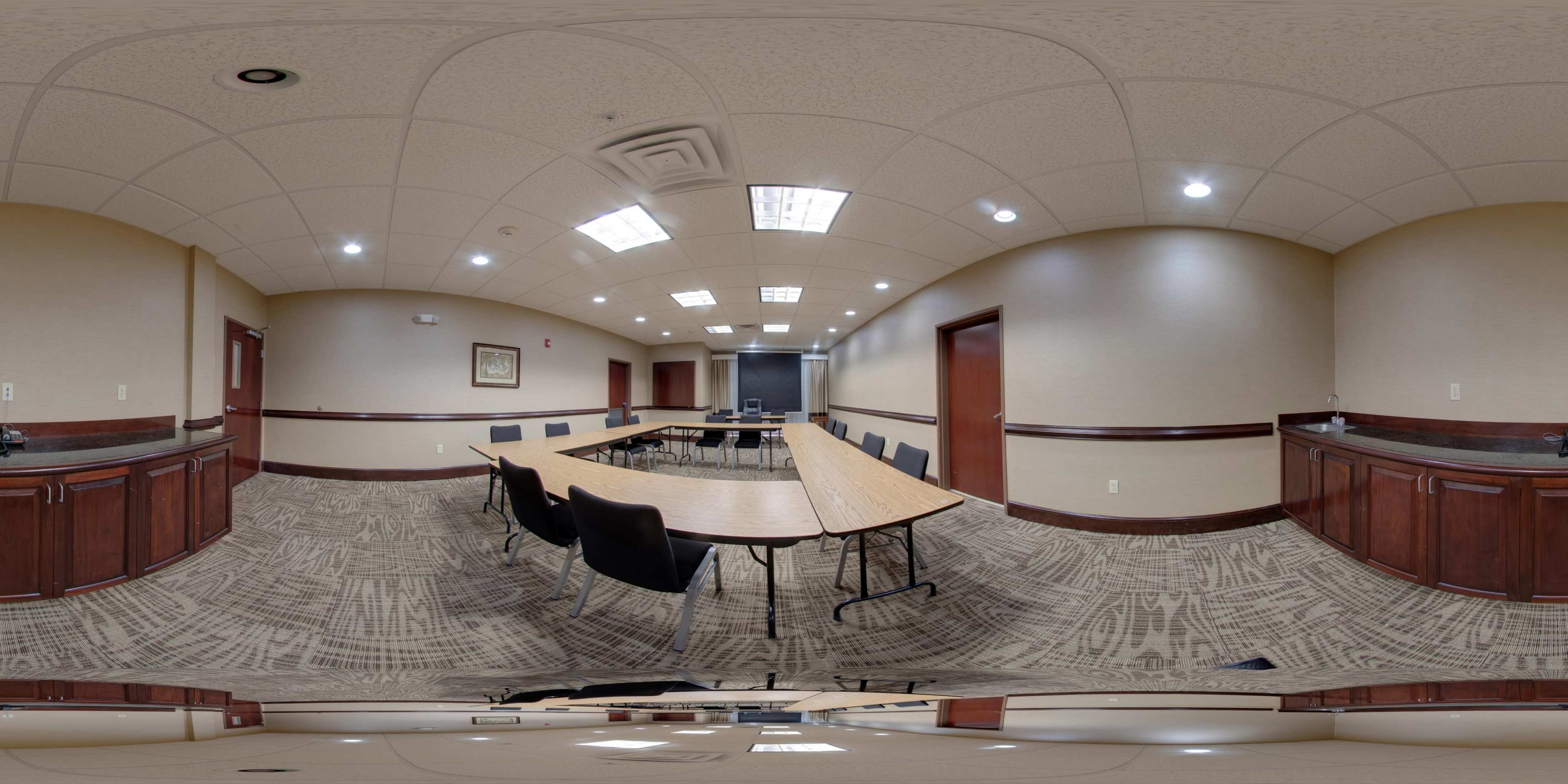 The image shows a conference room featuring a long table surrounded by black chairs and cabinets along the walls, with overhead lights illuminating the space.