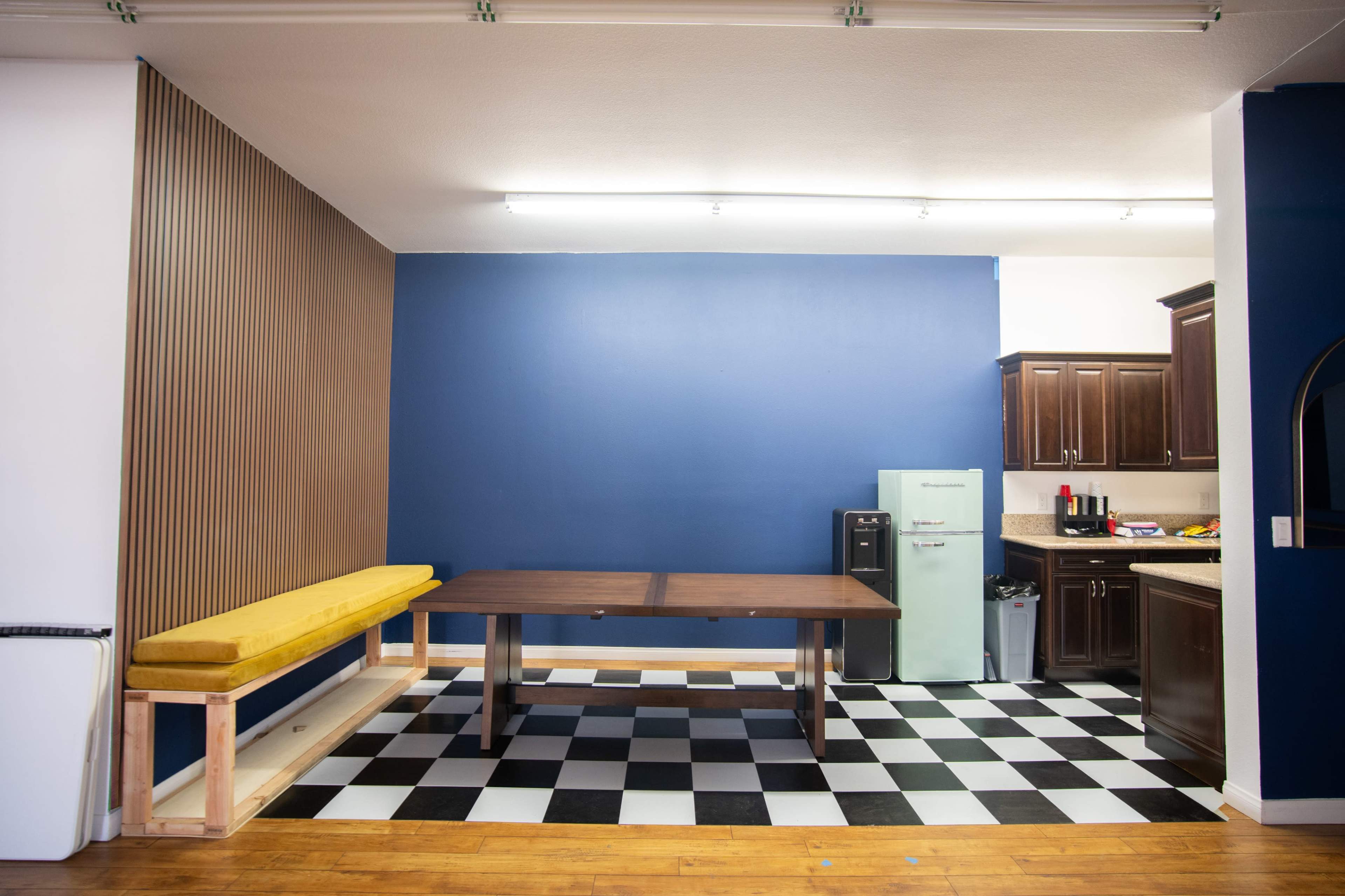 The image shows a room with a yellow bench against a wooden wall, a wooden table in the center, a black and white checkered floor, and a kitchen area with dark cabinets on the right side.