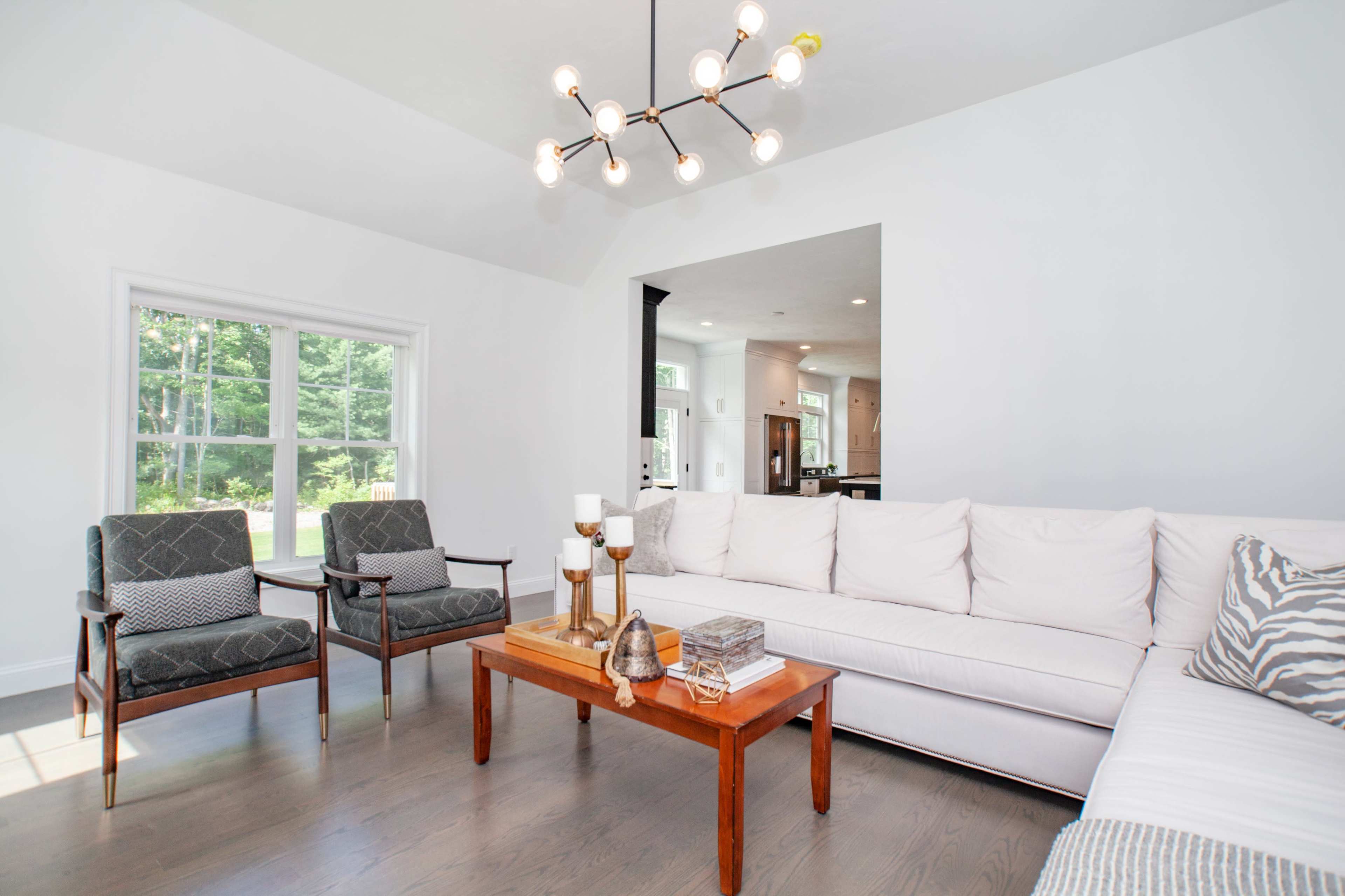 A well-lit living room featuring a white sectional sofa, two armchairs, a wooden coffee table, and a large window with a view of greenery outside.