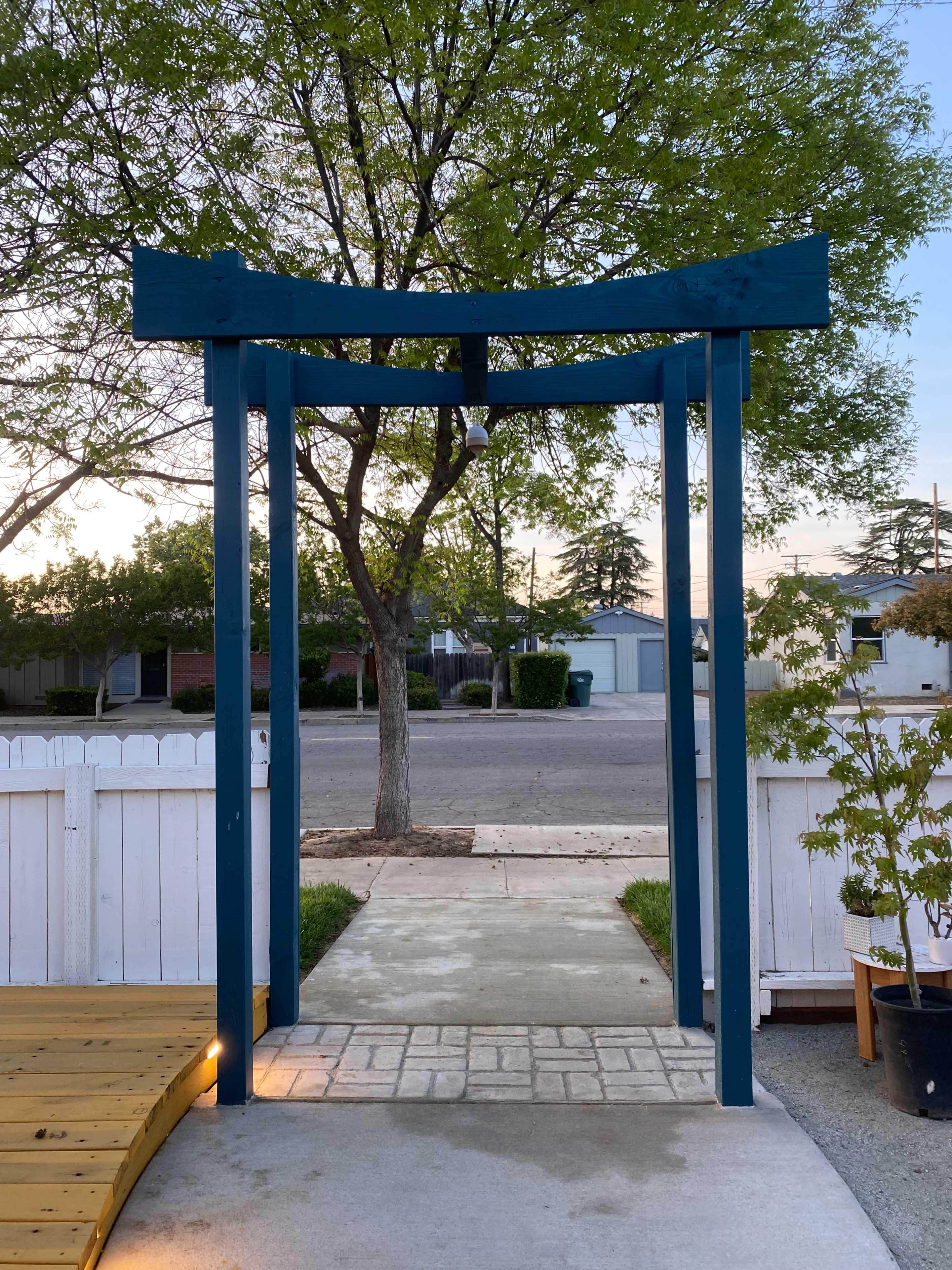 A blue torii gate leading to a stone pathway in a residential area with trees and houses in the background.