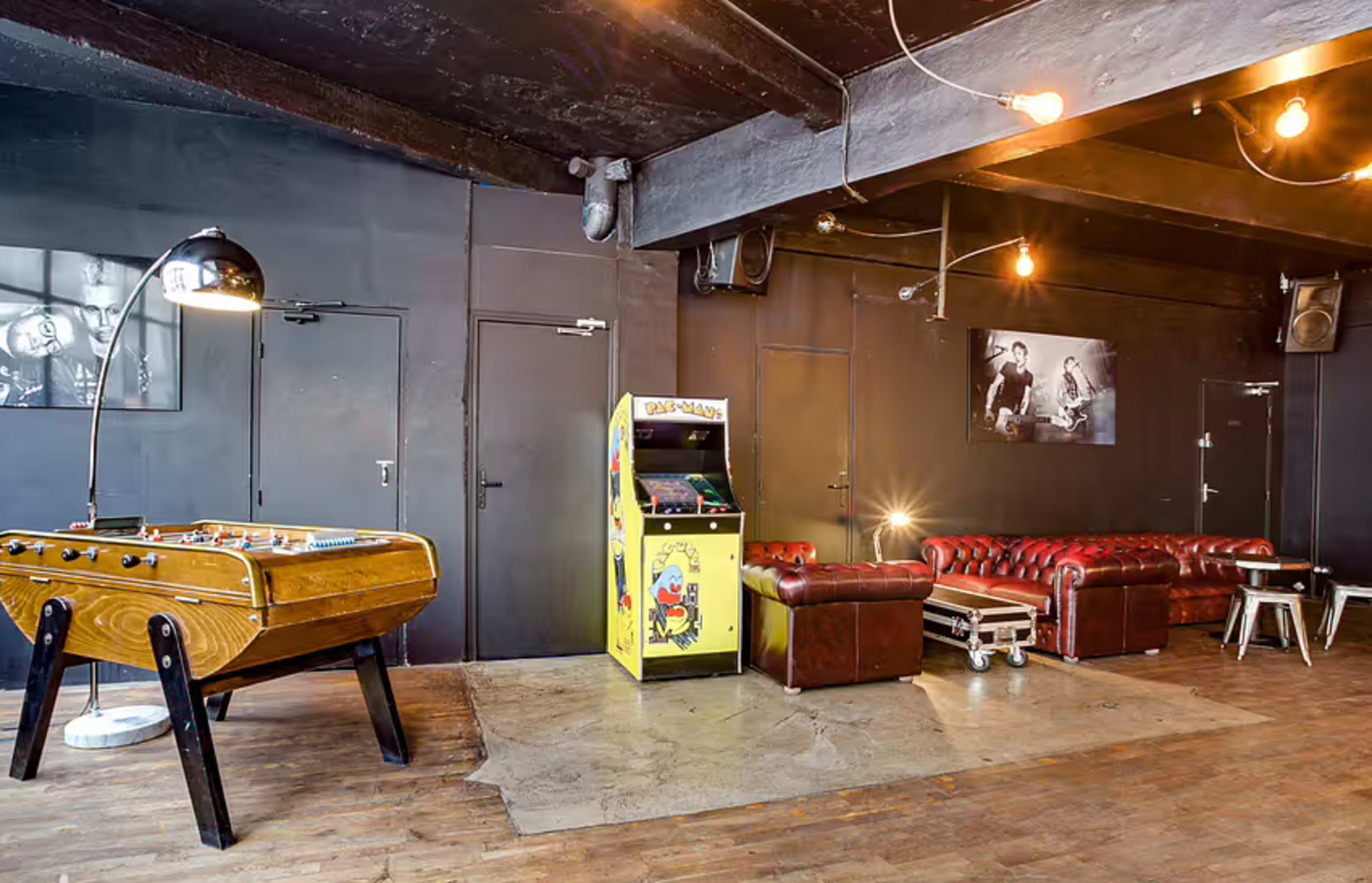 The image shows a dimly lit lounge area featuring a foosball table, a yellow arcade machine, a red leather couch, and industrial-style lighting.