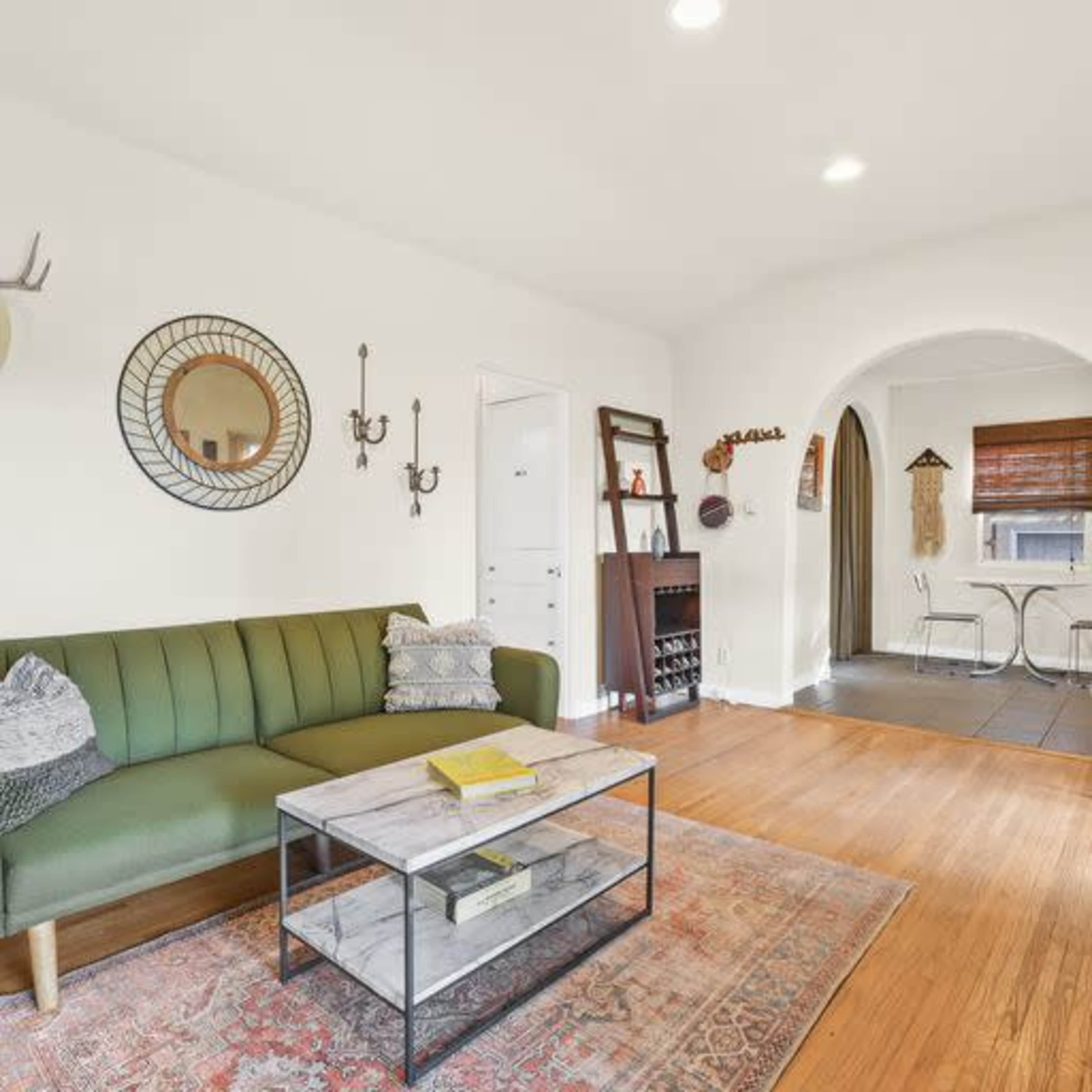 A living room with a green sofa, a round mirror on the wall, a coffee table, and a dining area in the background.