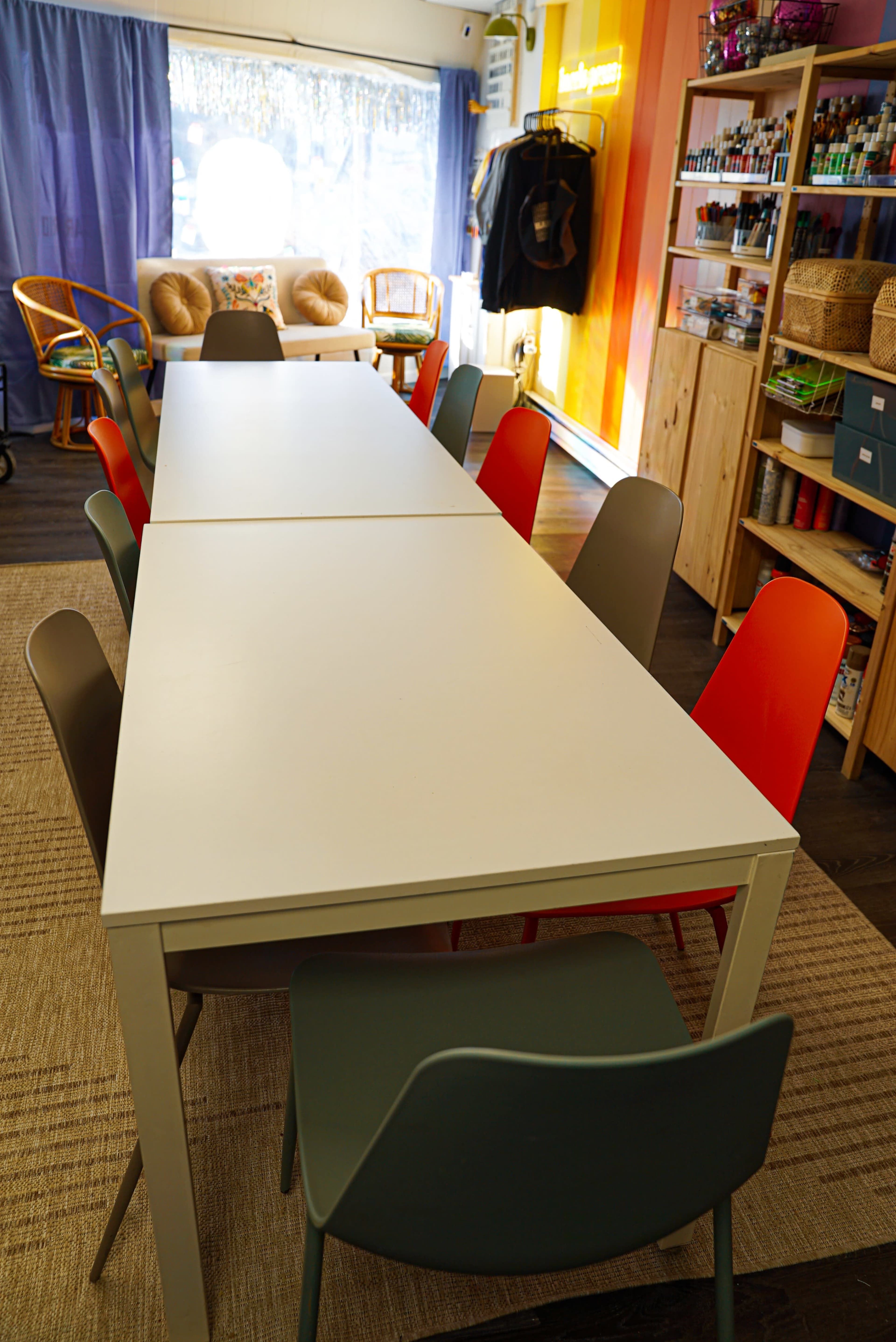 A long, rectangular white table surrounded by colorful chairs is set up in a bright room featuring shelves filled with various items and a window with natural light.