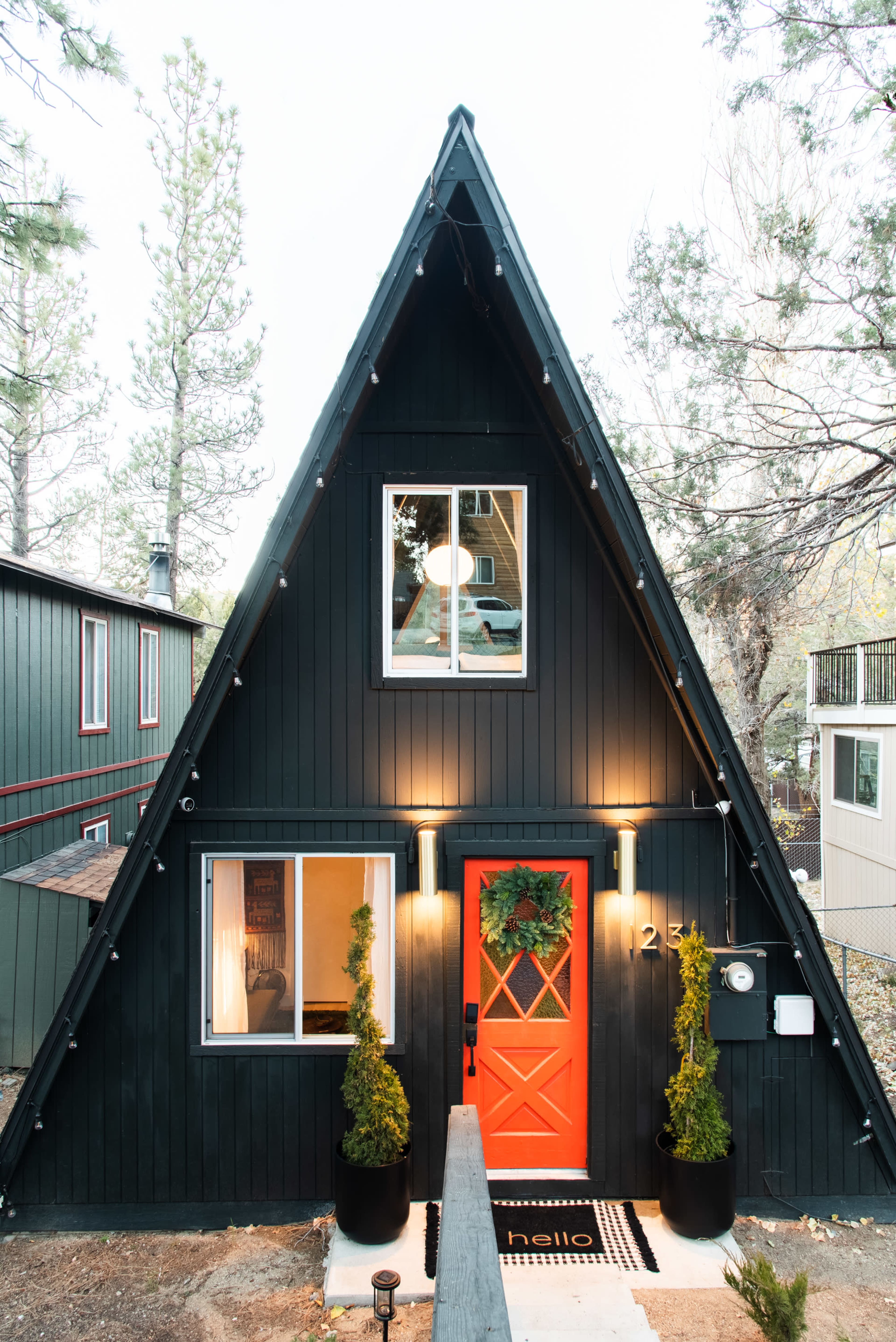 A black A-frame house with a bright orange door and two small evergreen shrubs on either side, surrounded by tall pine trees.