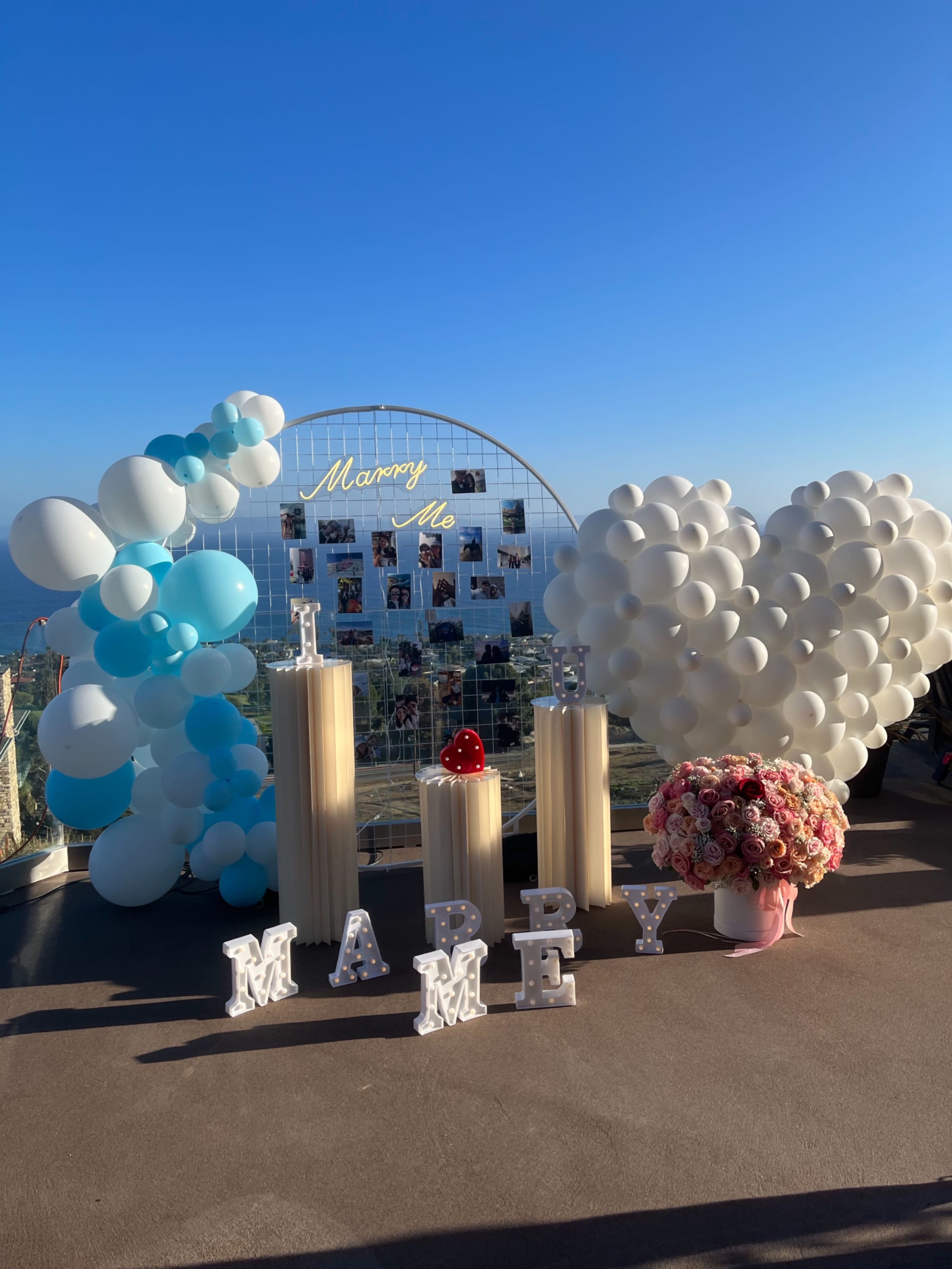 A decorative setup for a marriage proposal, with balloons in blue and white, floral arrangements, and a sign that reads "Marry Me" surrounded by photo displays.