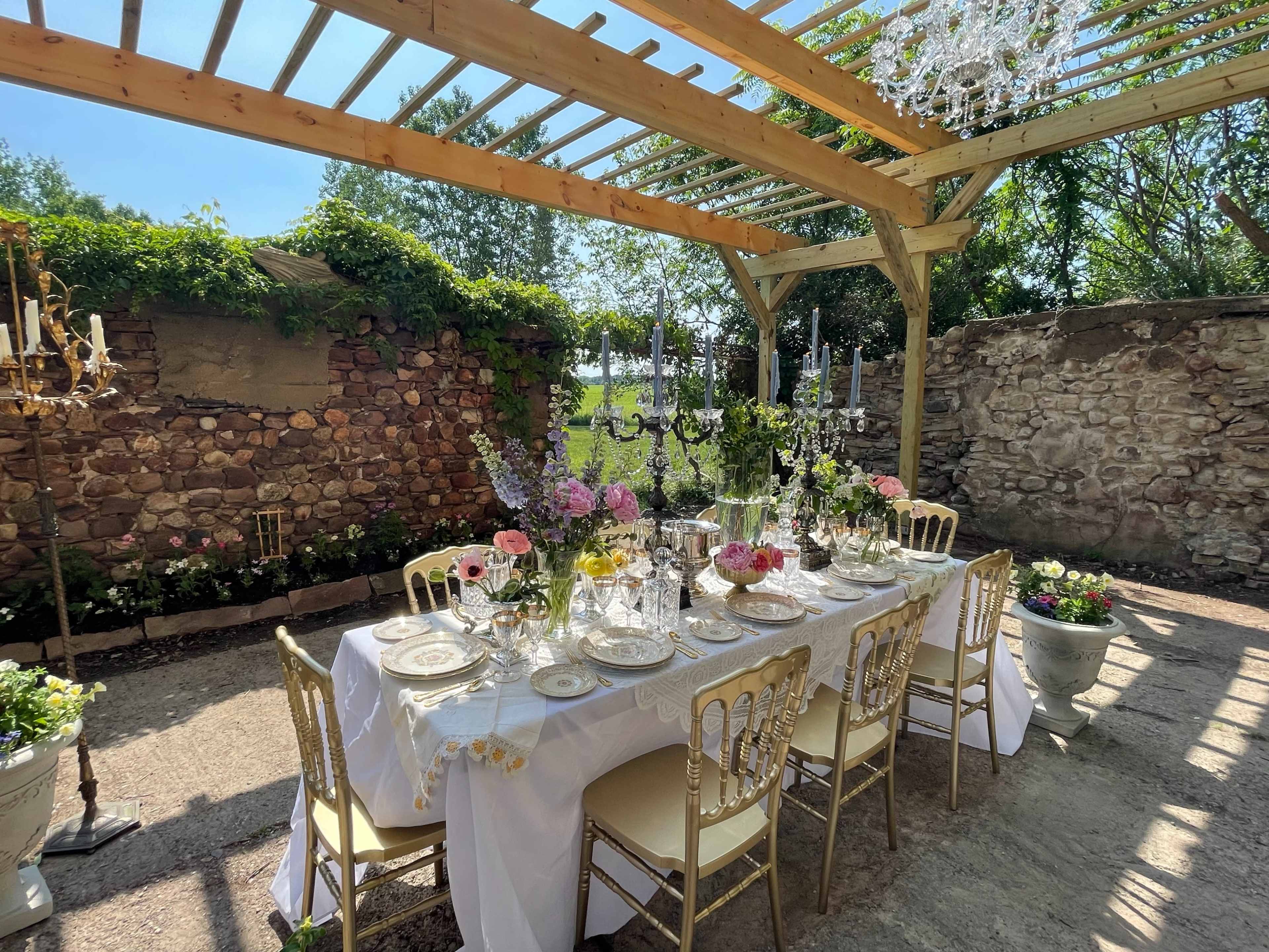 A long dining table set with elegant tableware and floral arrangements is positioned under a wooden pergola in a garden space.