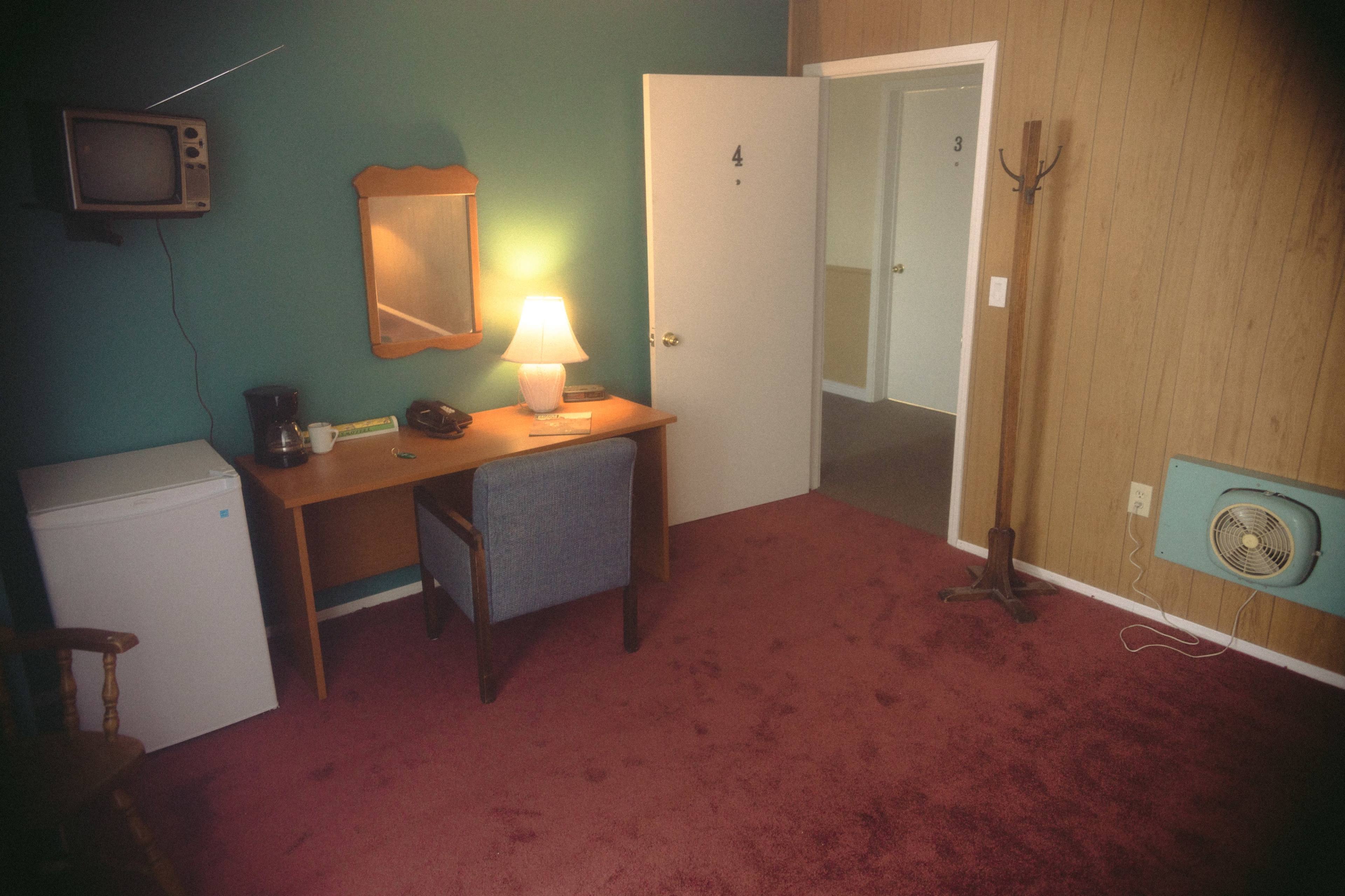 A hotel room features a desk with a chair, a lamp, a mirror, a mini fridge, and a television mounted on the wall, with an open door leading to another room.
