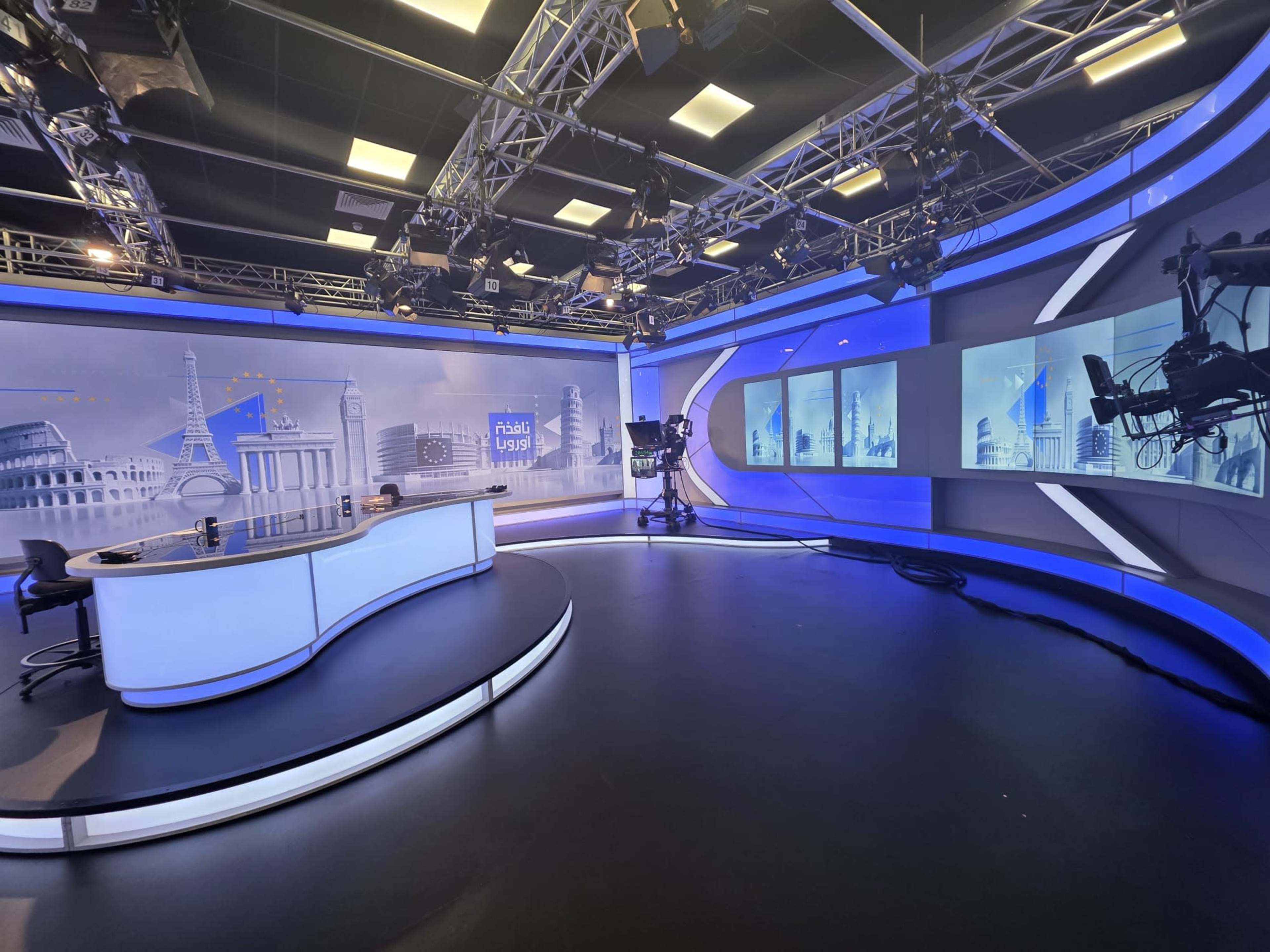 The image shows a modern television studio with a curved news desk, a camera setup, and large screens displaying a cityscape background.