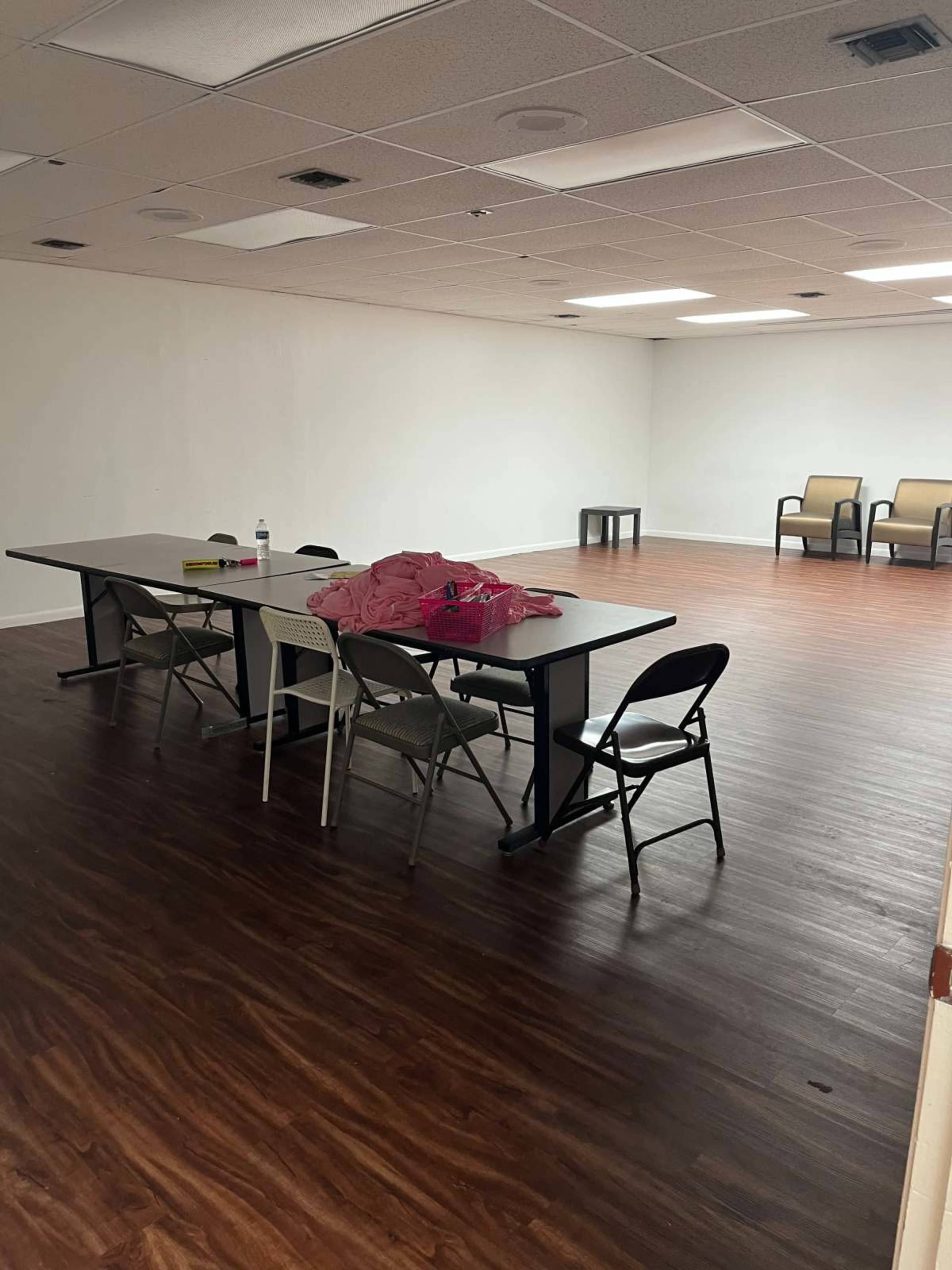 The image shows a sparse room with a table and several chairs arranged near the center, and a few empty chairs along one wall.
