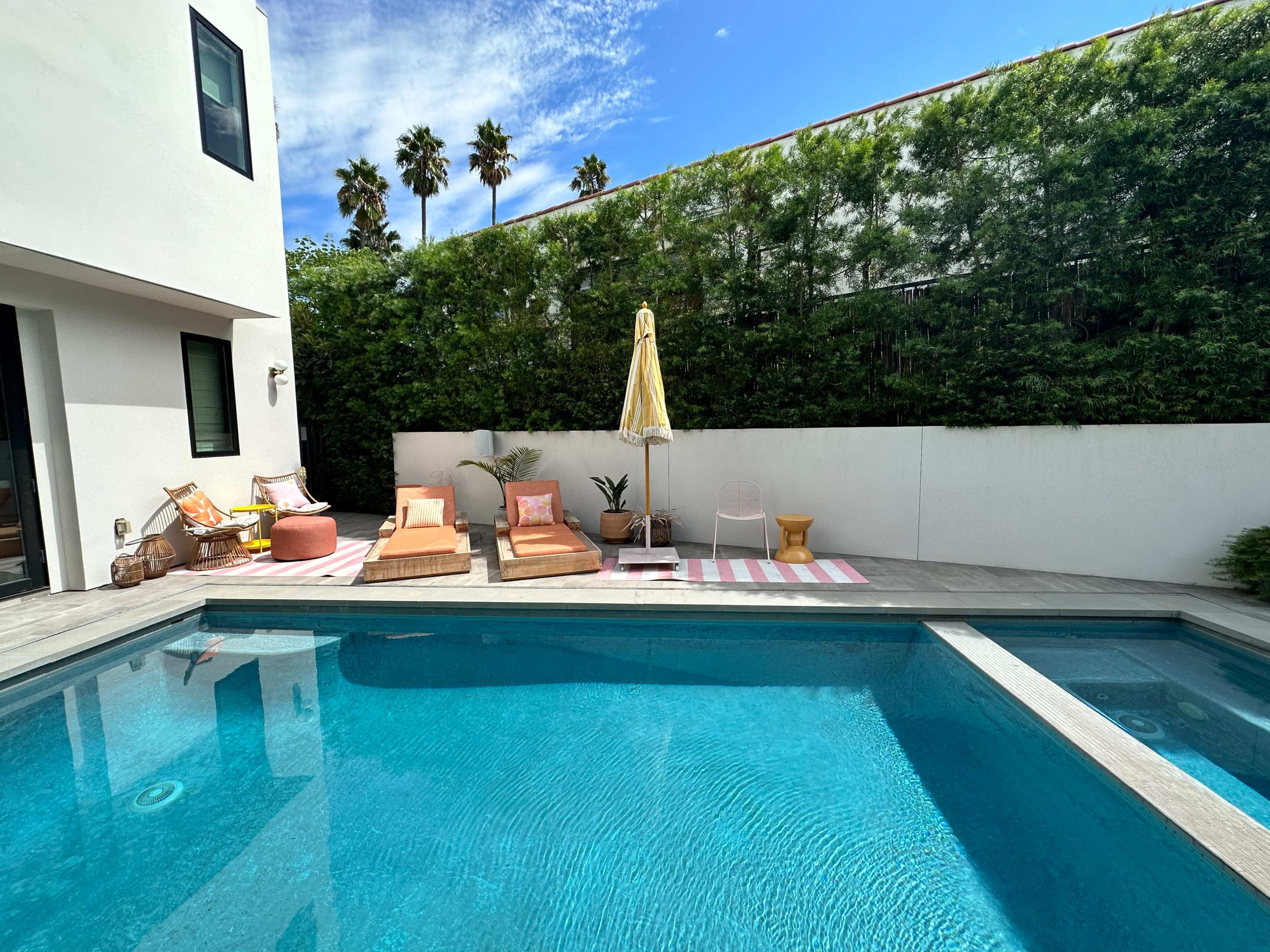 A modern pool area with loungers, an umbrella, and greenery surrounding a clear pool under a blue sky.