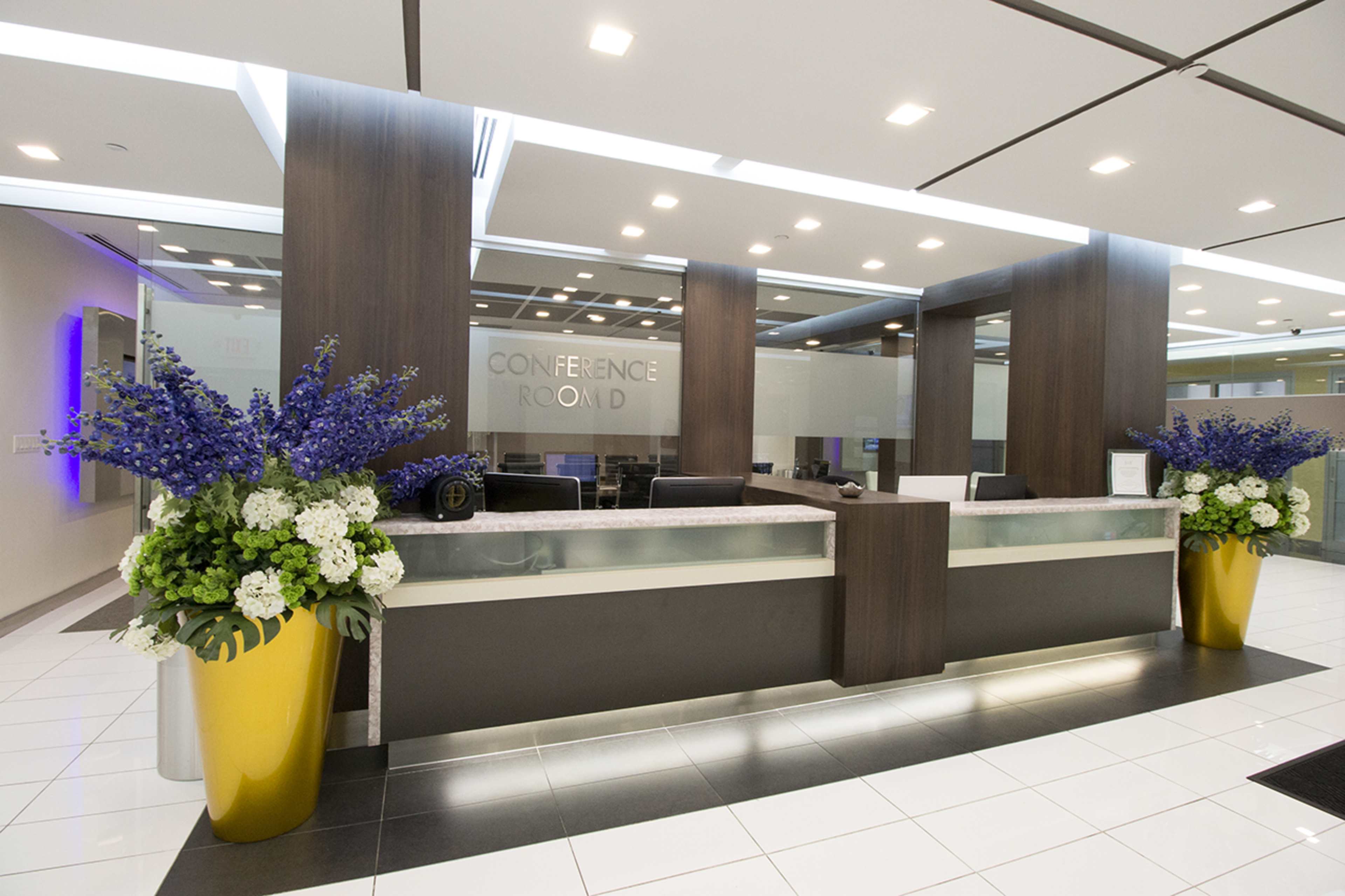 The image shows a modern reception area for a conference room, featuring a desk with floral arrangements on either side and clear signage indicating "CONFERENCE ROOM."