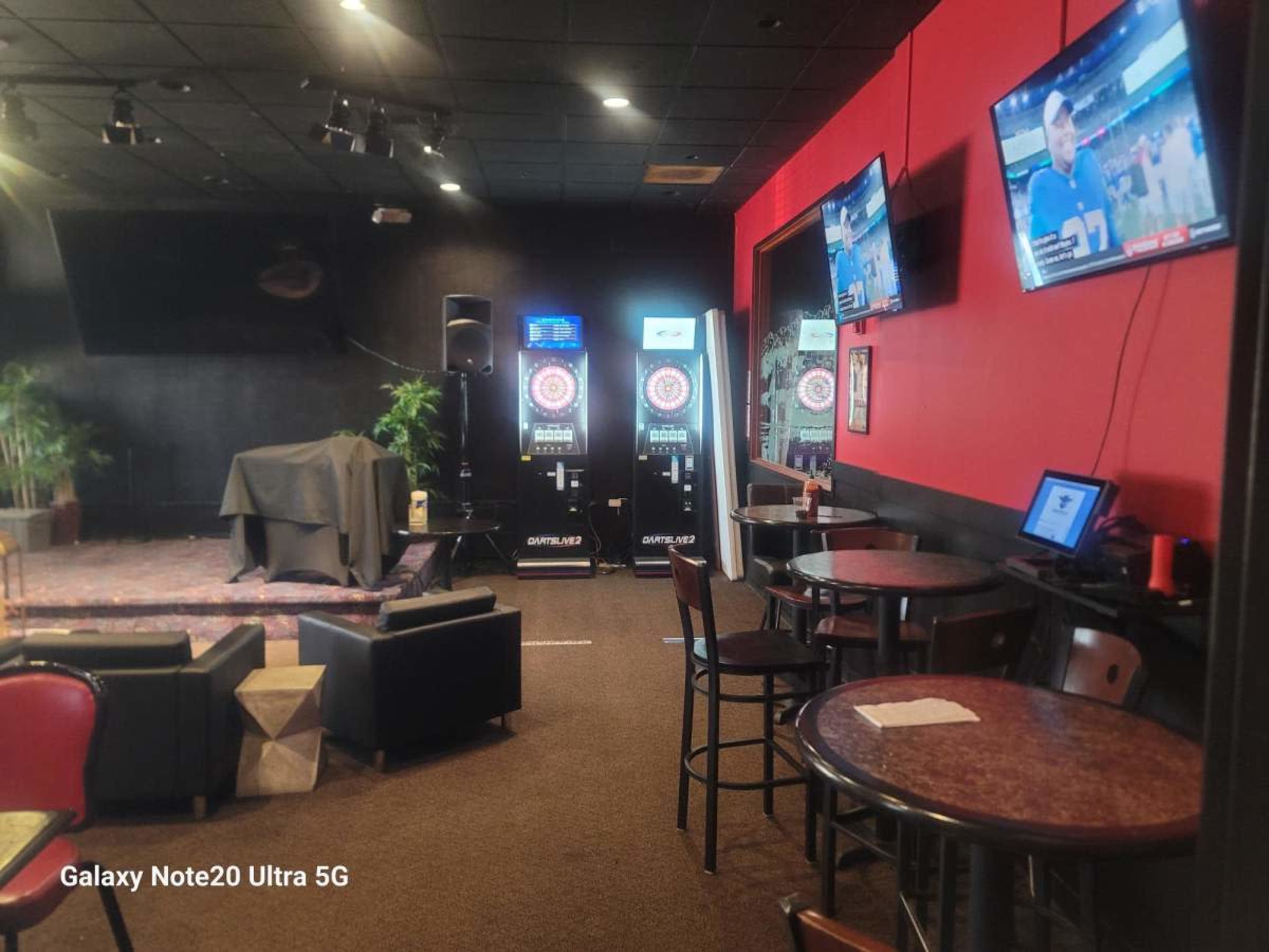 The image shows a bar area with several tables and chairs, two screens displaying a sports game, and dart machines in the background.