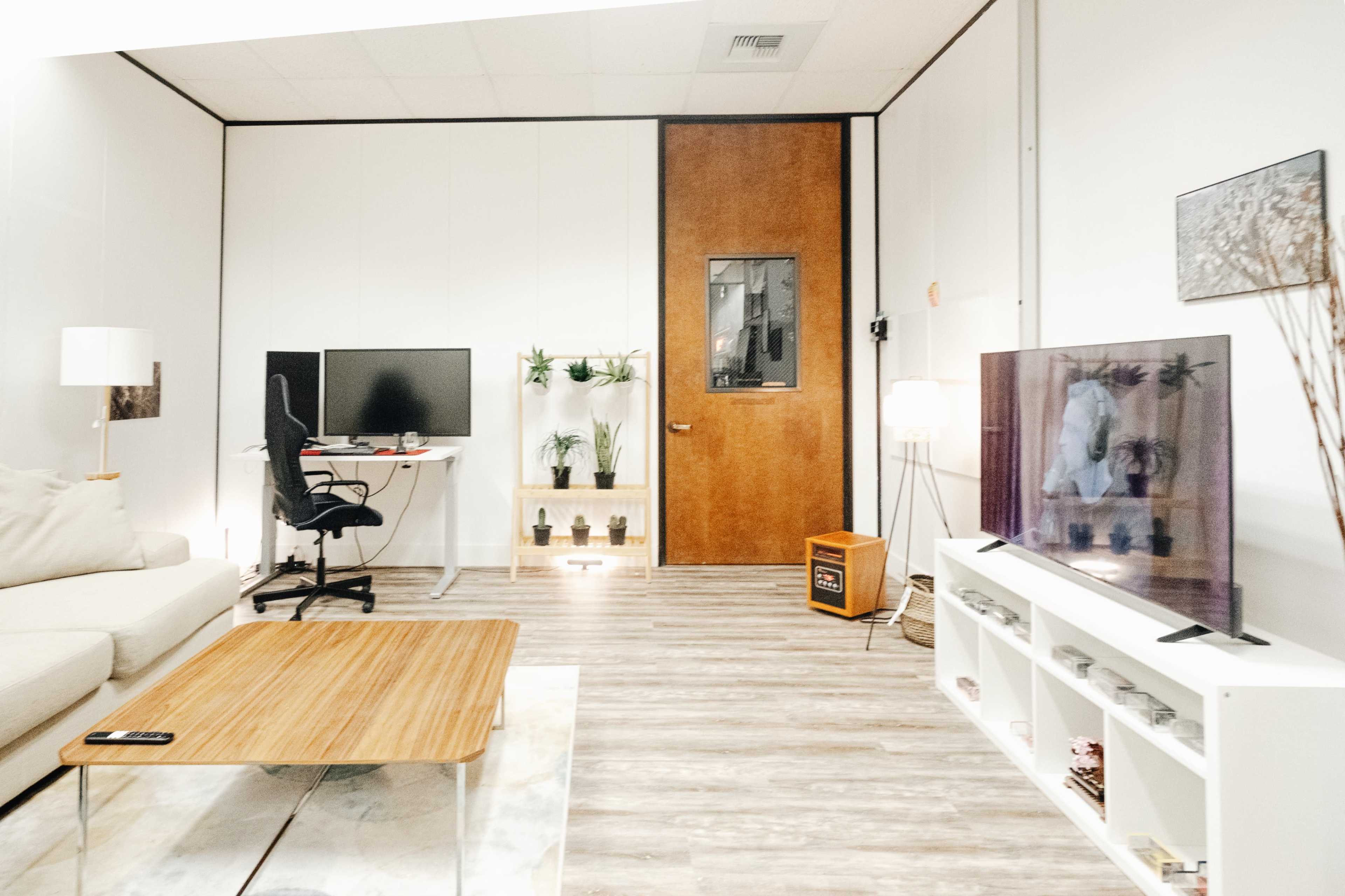 A modern, minimalistic workspace featuring a desk with a monitor, a chair, a coffee table, and decorative plants, against a backdrop of a wooden door and a television.