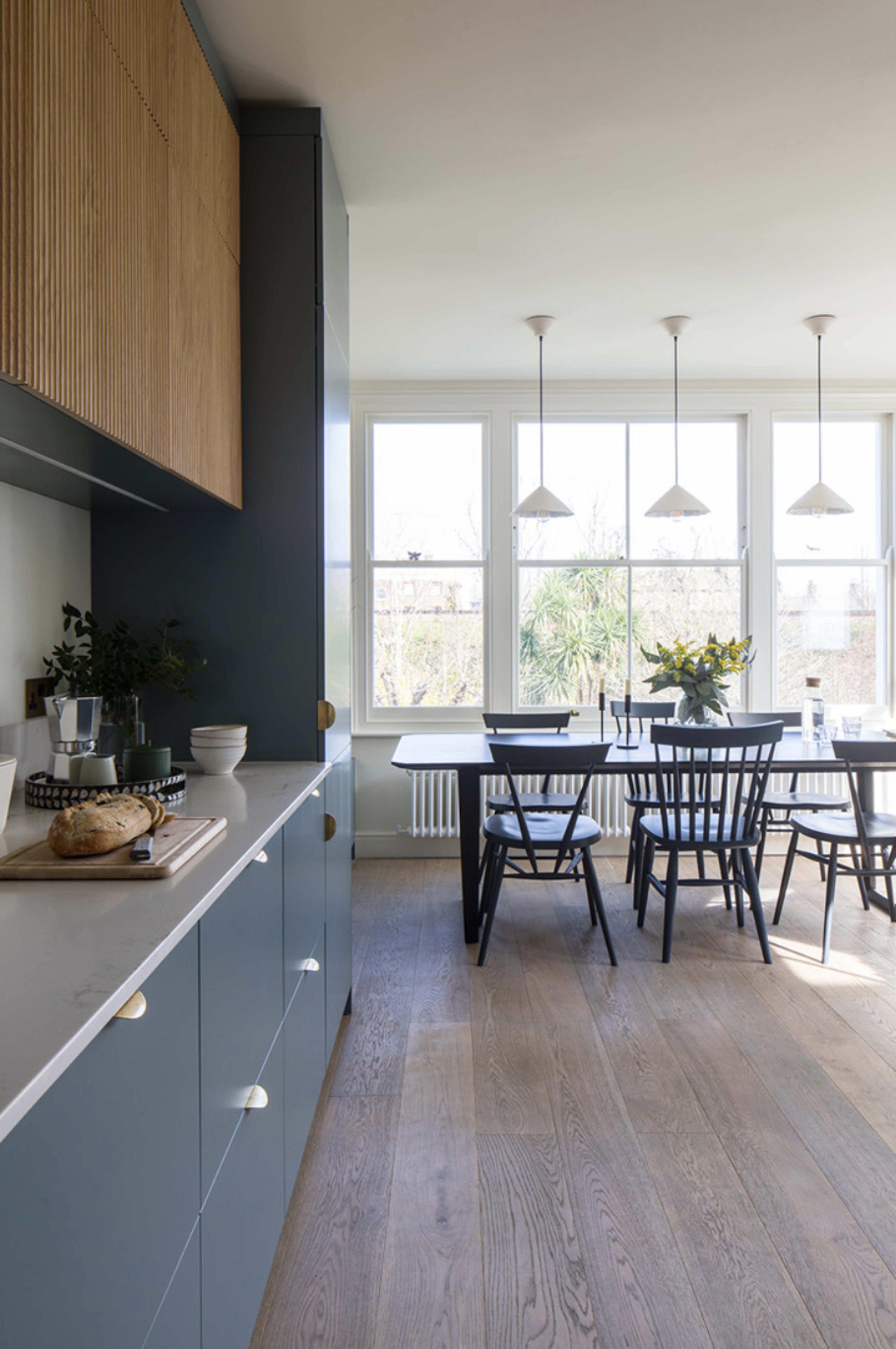 A modern kitchen with blue and wooden cabinetry, featuring a dining area with a black table and chairs illuminated by pendant lights.