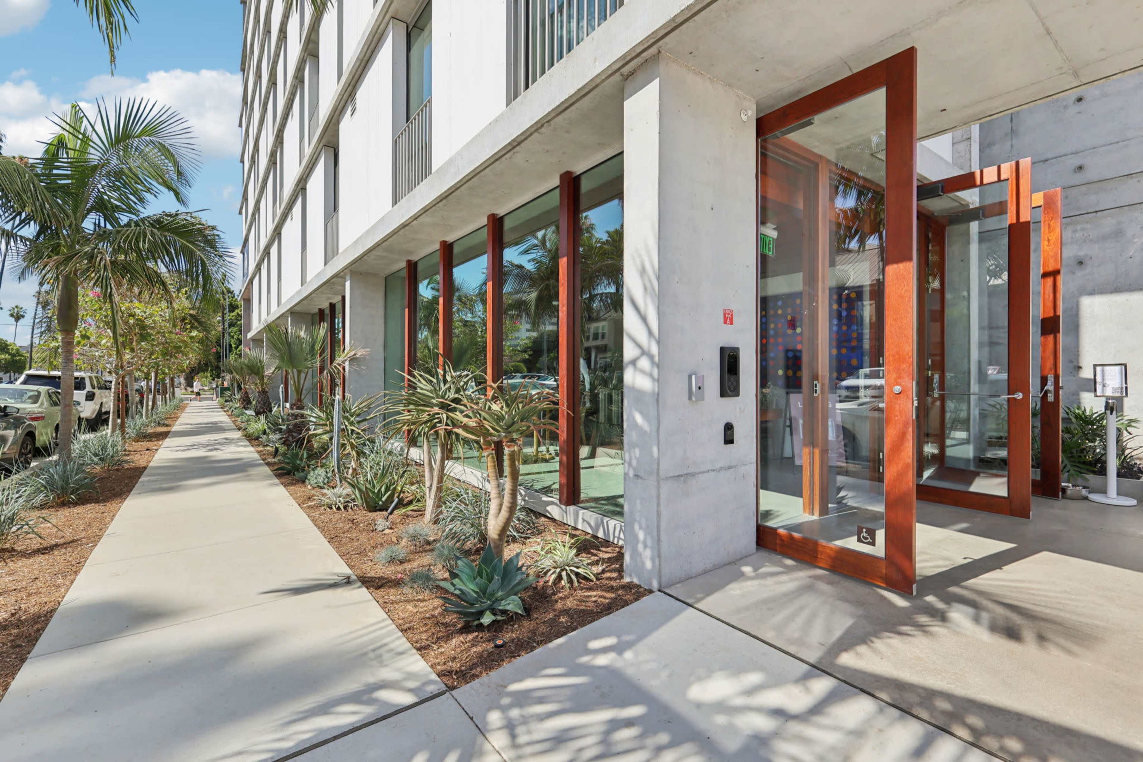 The image shows a modern building entrance with glass doors surrounded by landscaped greenery and a concrete walkway.