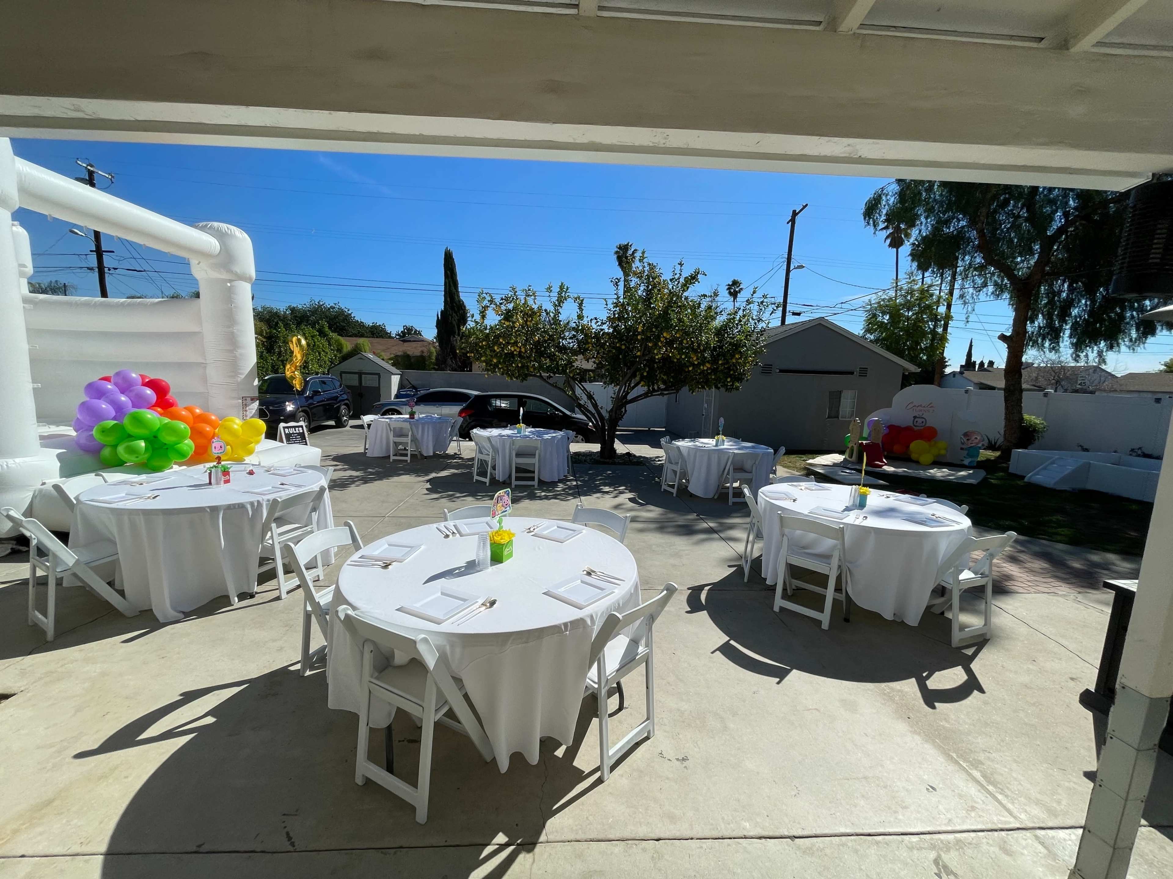 The scene shows a sunlit outdoor area set up for a party, featuring whiteTables with table settings and colorful balloons in the background.