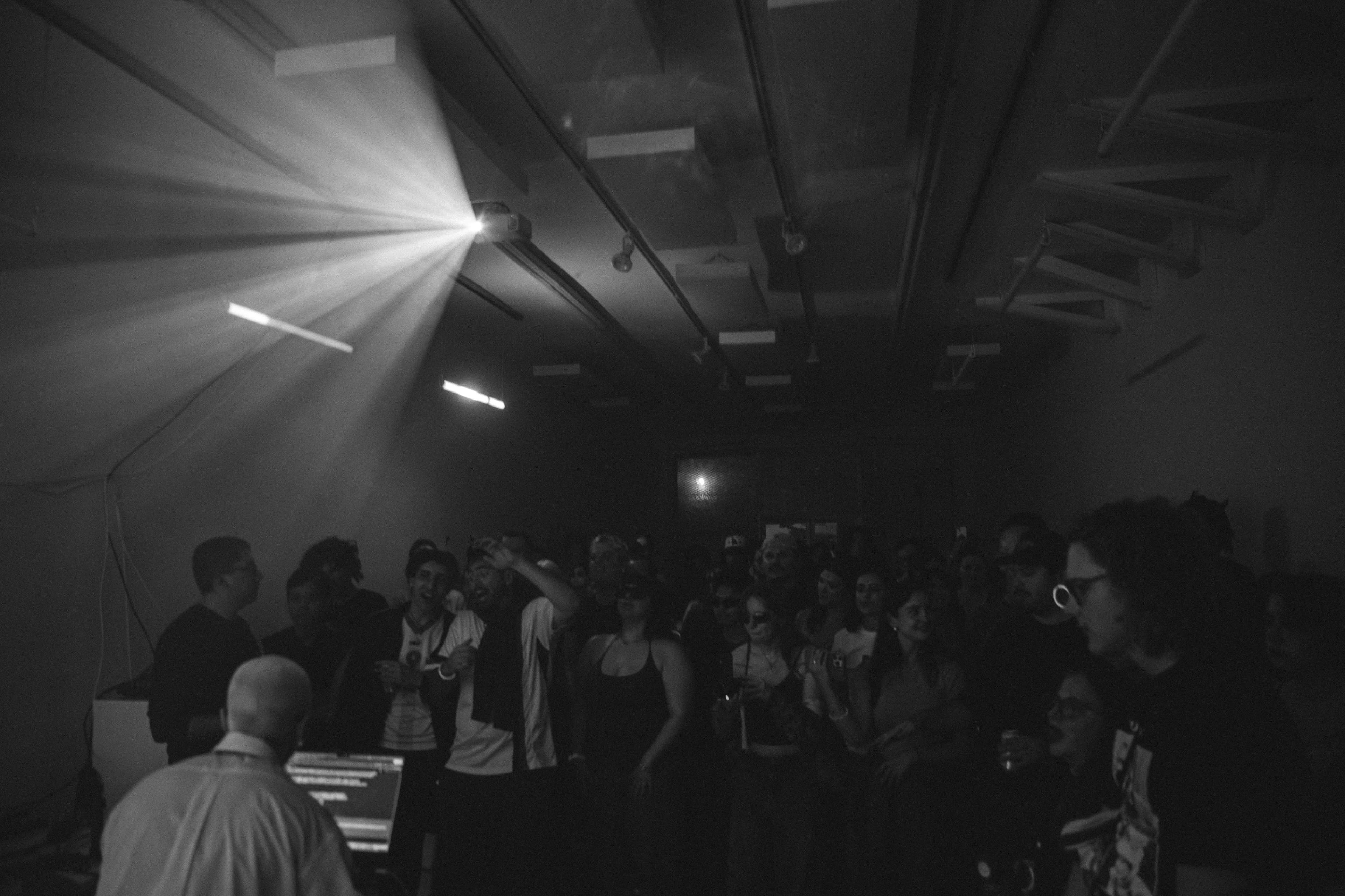 A crowd of people attentively watches a performer in a dimly lit room, with beams of light illuminating the space.