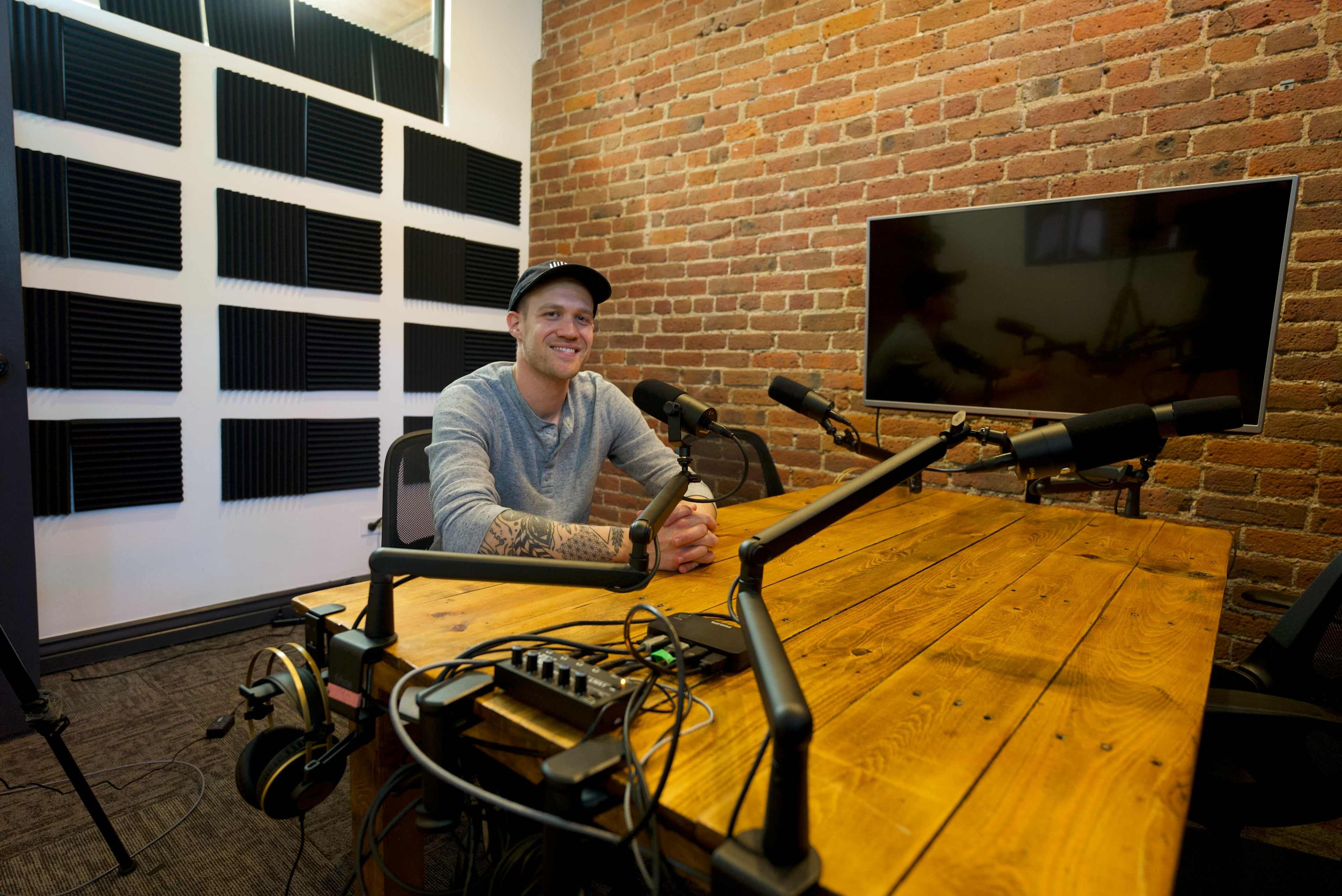 A man sits at a wooden table in a soundproof recording studio with brick walls and audio equipment visible.
