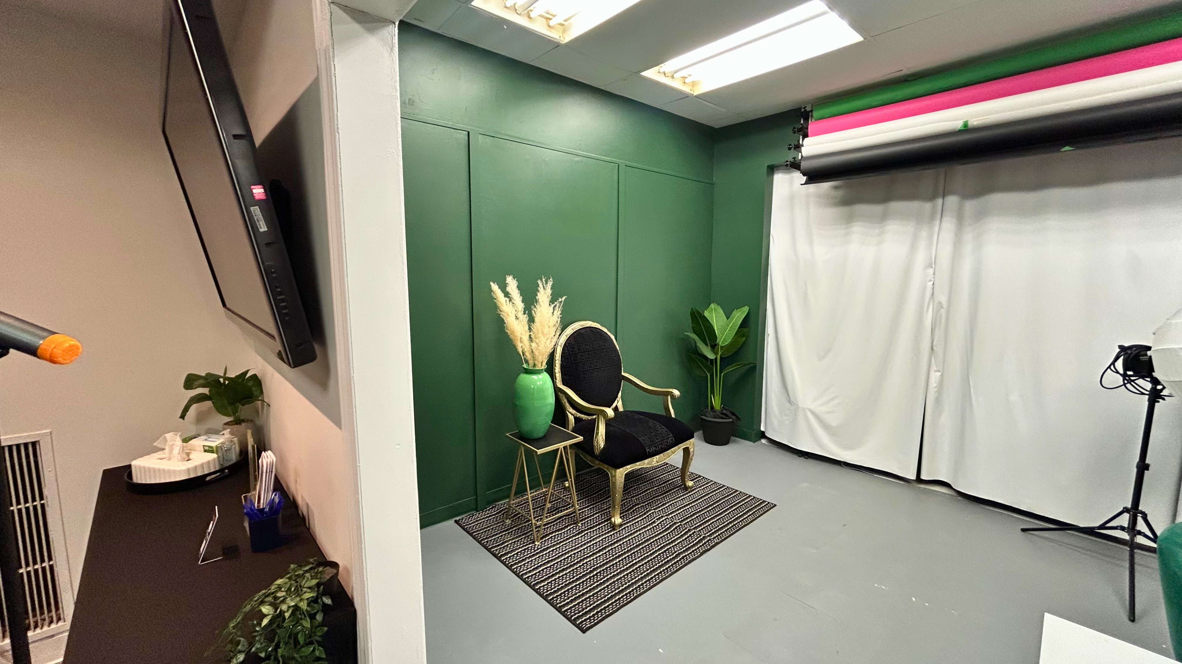 The image shows a small studio space featuring a black chair with gold accents, a green vase with dried plants, a potted plant, and a backdrop setup with a white curtain and colorful lighting.