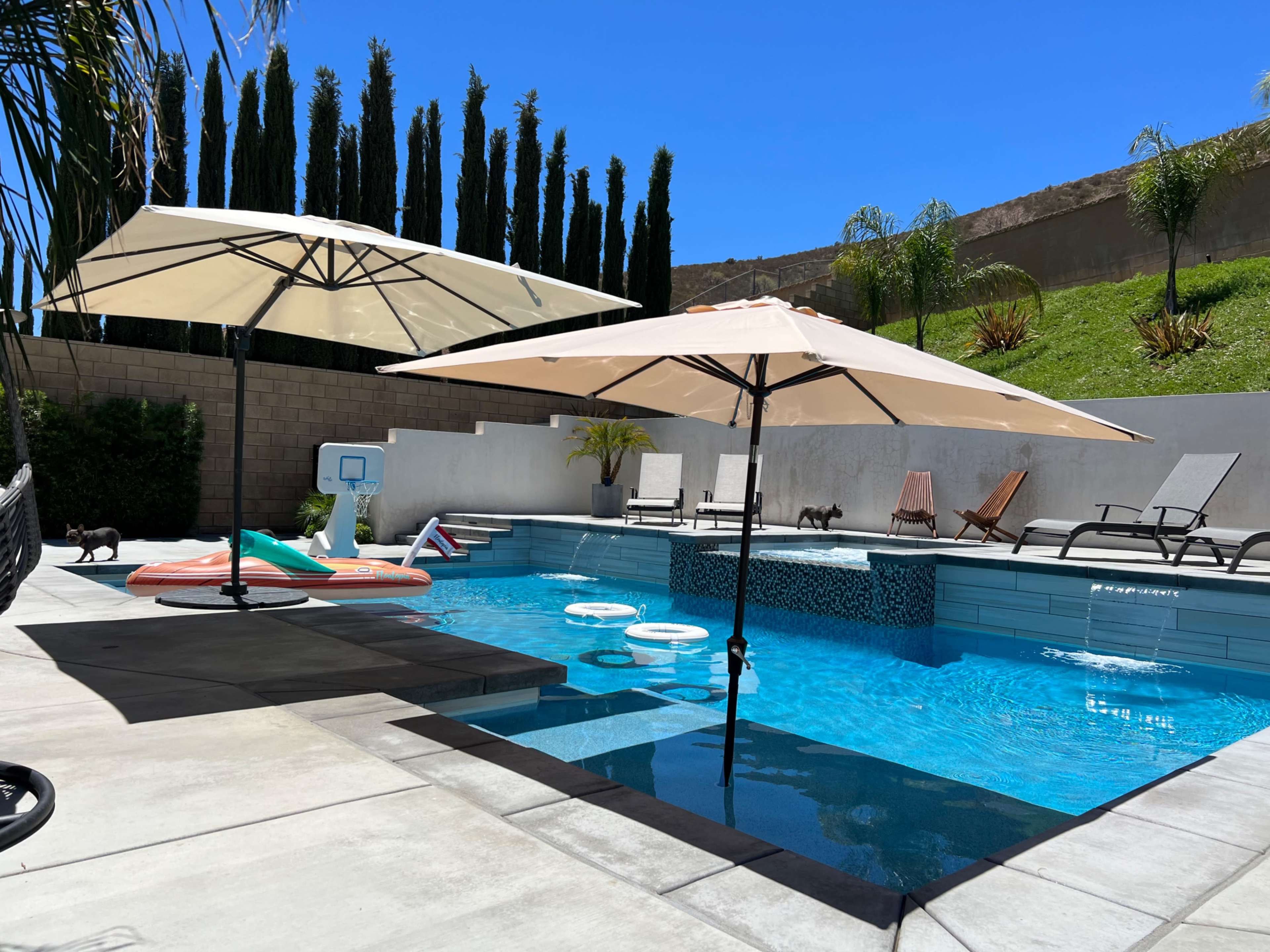 A swimming pool area with two large umbrellas, lounge chairs, a basketball hoop, and inflatable pool toys, surrounded by tall, green trees and a blue sky.