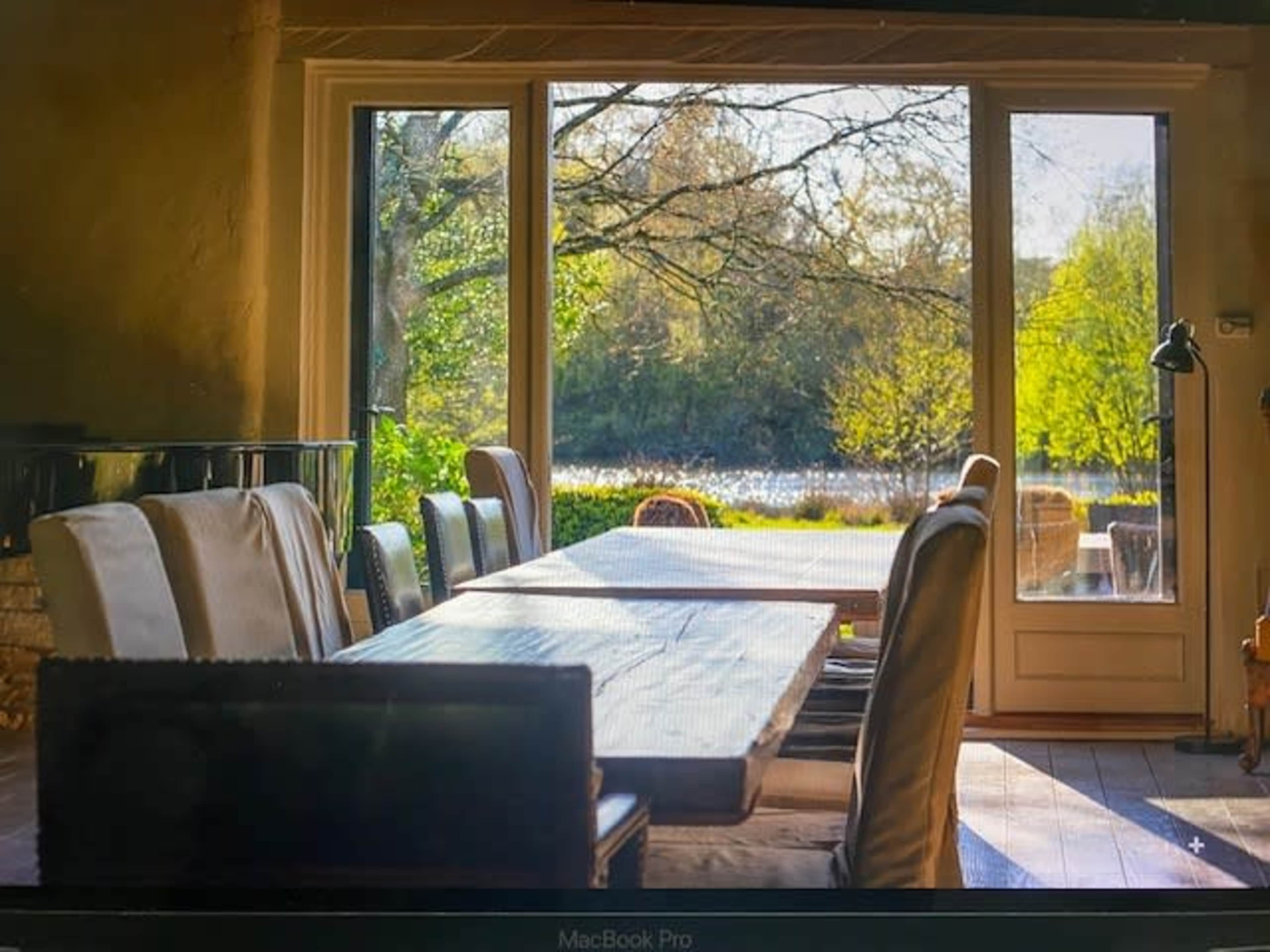 A large wooden dining table with chairs is visible in a room, overlooking a lake and trees through large glass doors.