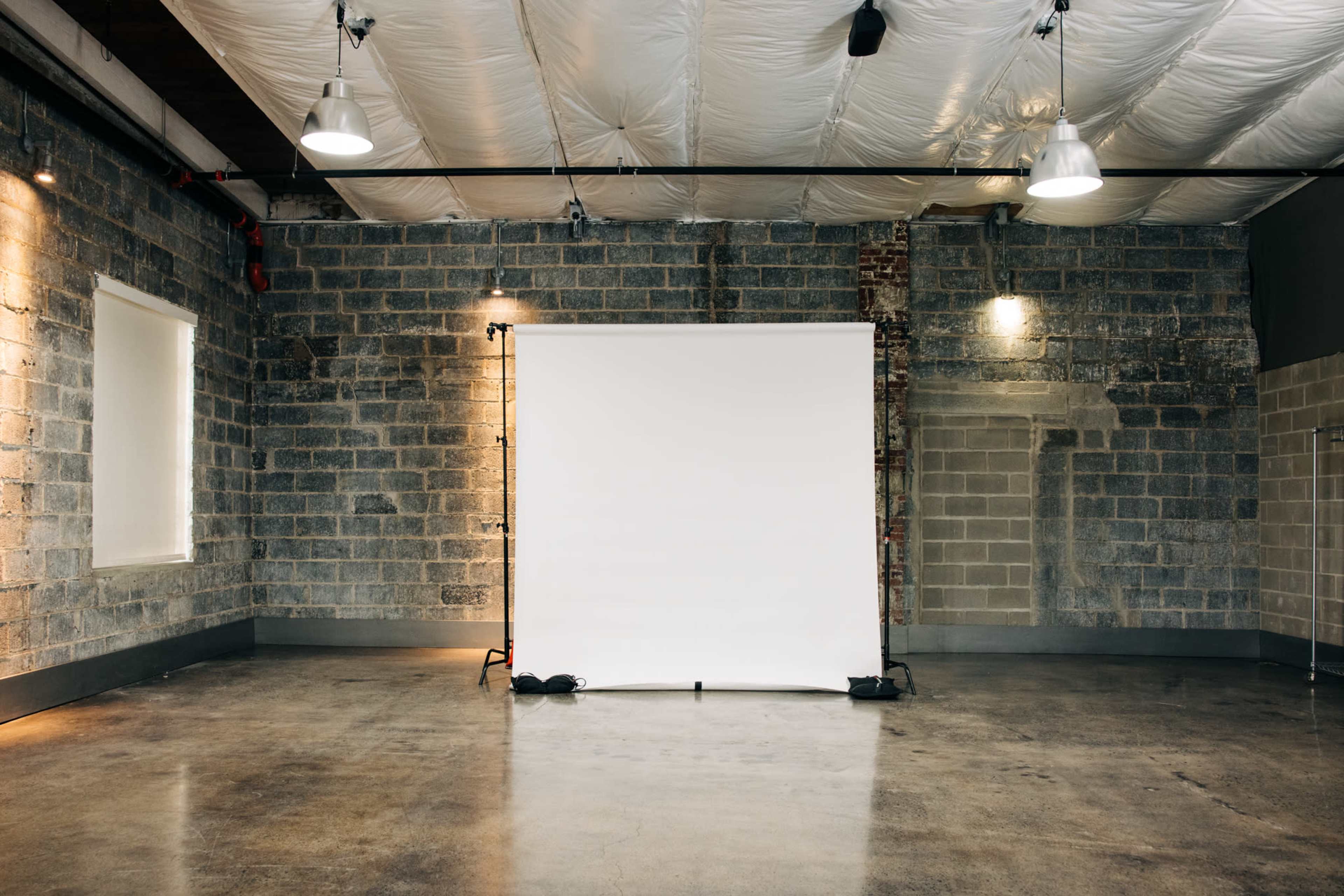 The image shows an empty industrial-style room with exposed brick walls and a large white backdrop in the center.