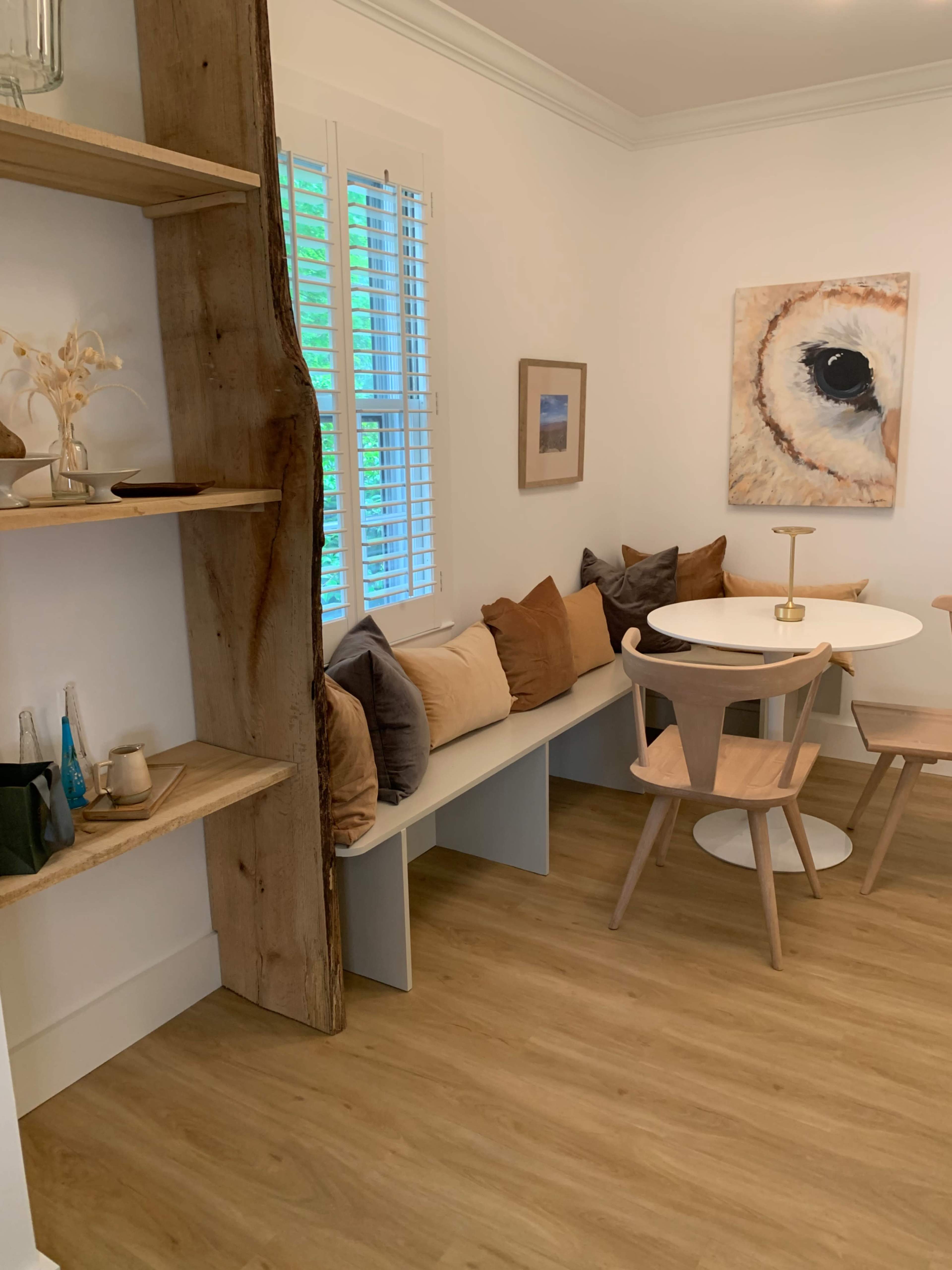 A cozy dining area features a circular table surrounded by wooden chairs and a built-in bench with cushions, beside a wooden shelf and a large owl painting.