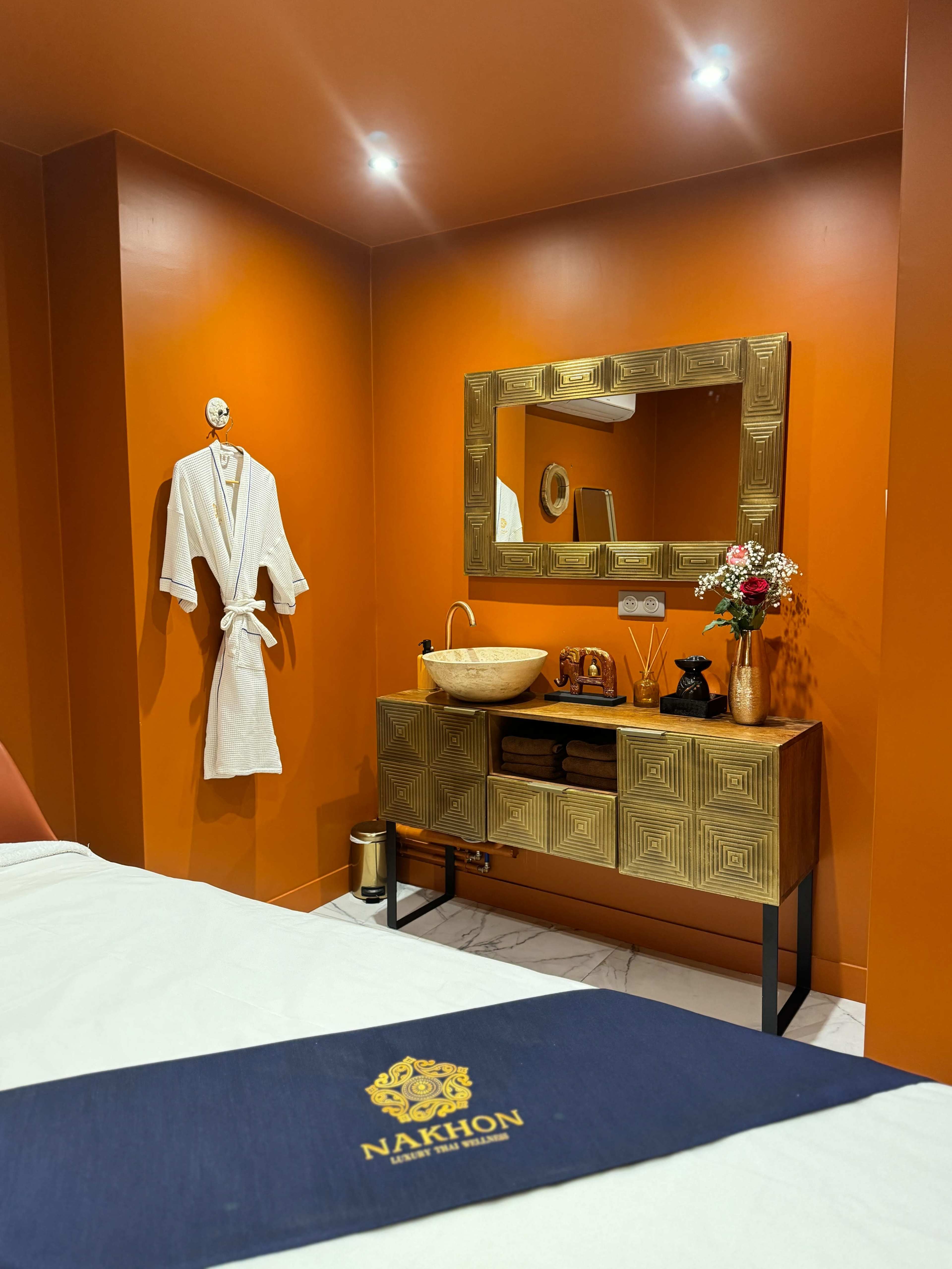The image shows a well-decorated spa room with an orange wall, a gold-framed mirror, a sink on a wooden console, and a bathrobe hanging nearby.