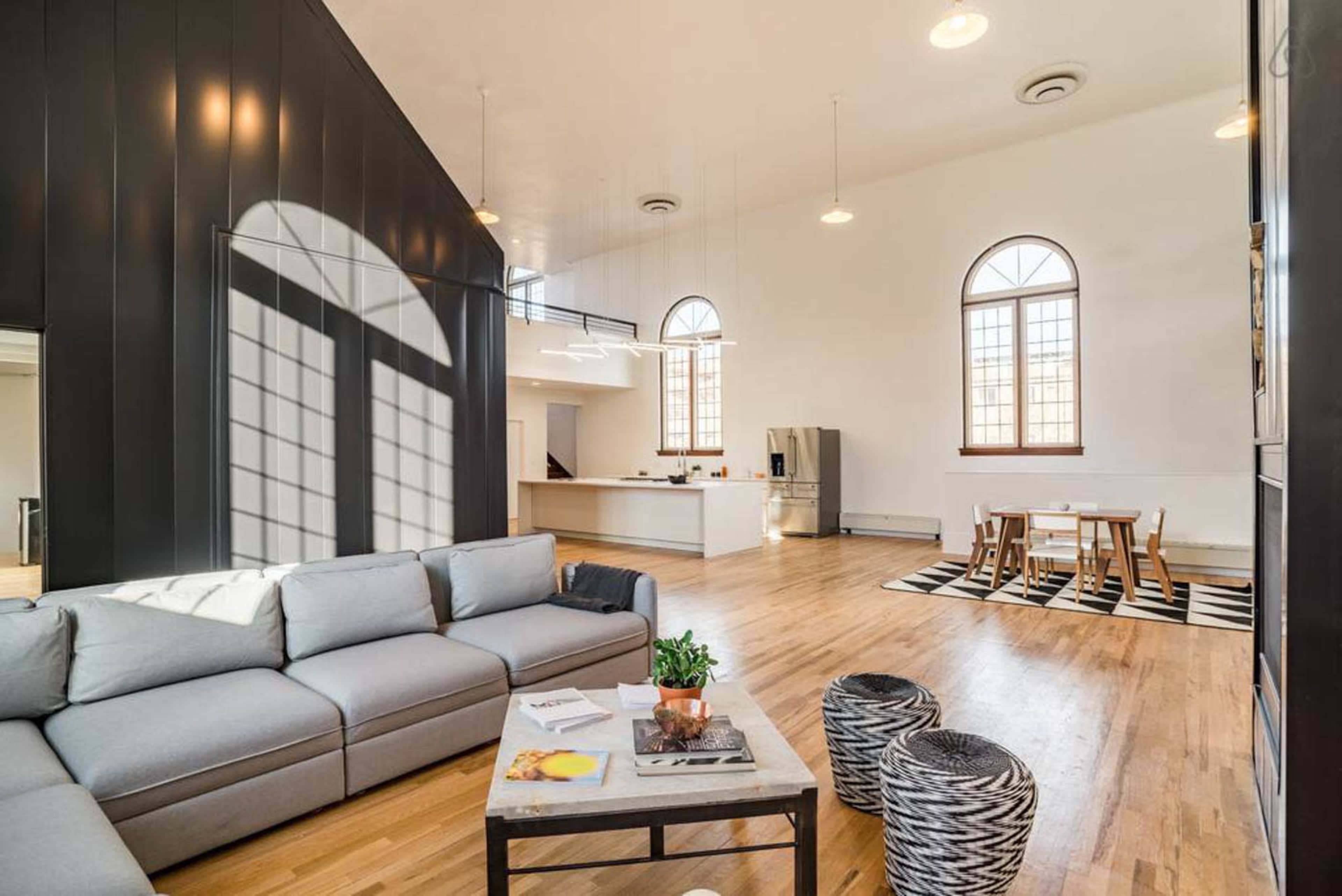 The image shows a spacious, modern living area with a large gray sofa, a wooden dining table, and a minimalist kitchen in the background.