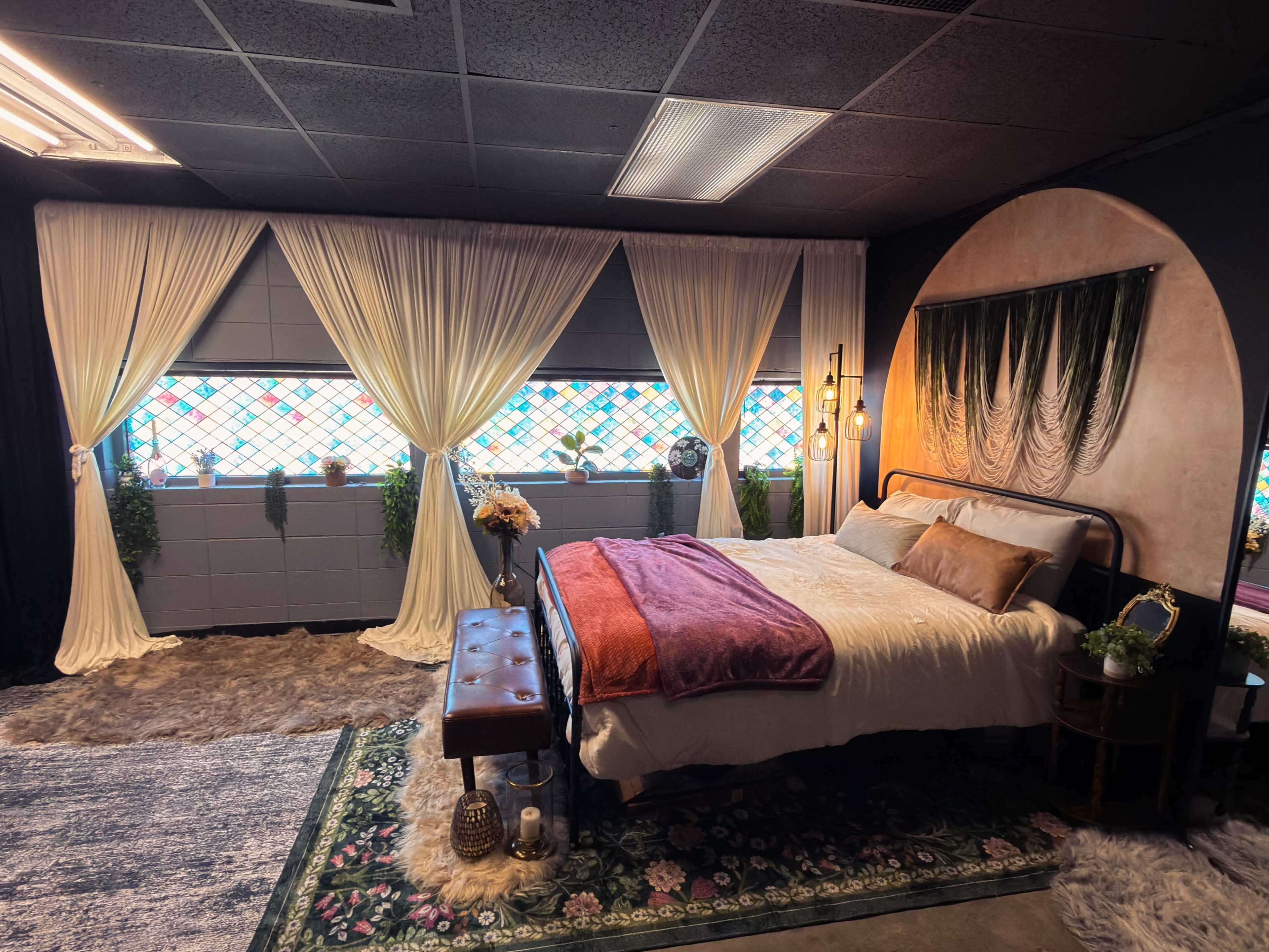 The image shows a bedroom featuring a bed with white linens, decorative curtains, stained glass windows, and various plants and furnishings.