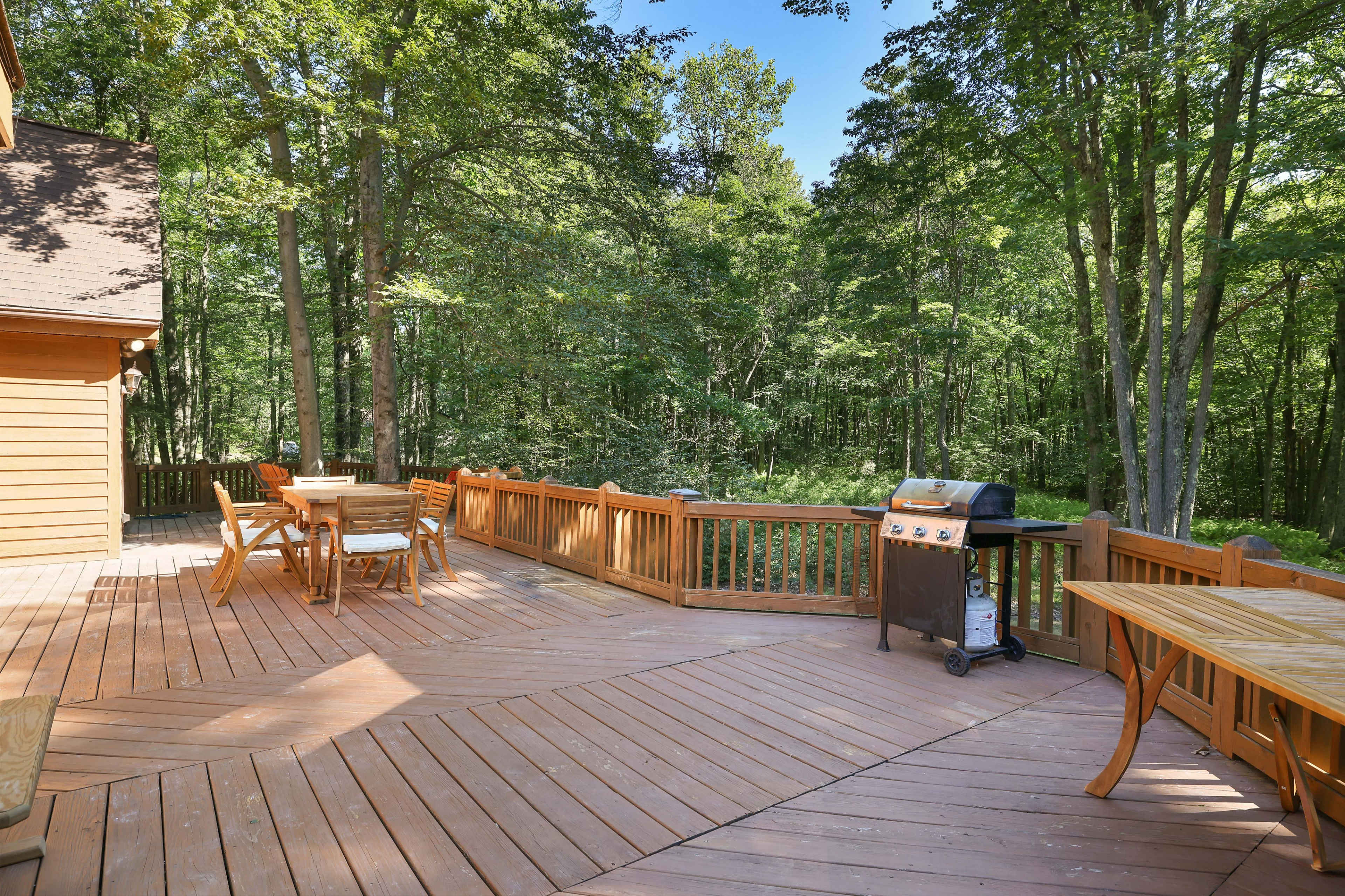 A wooden deck features a grill and several outdoor dining tables surrounded by trees.