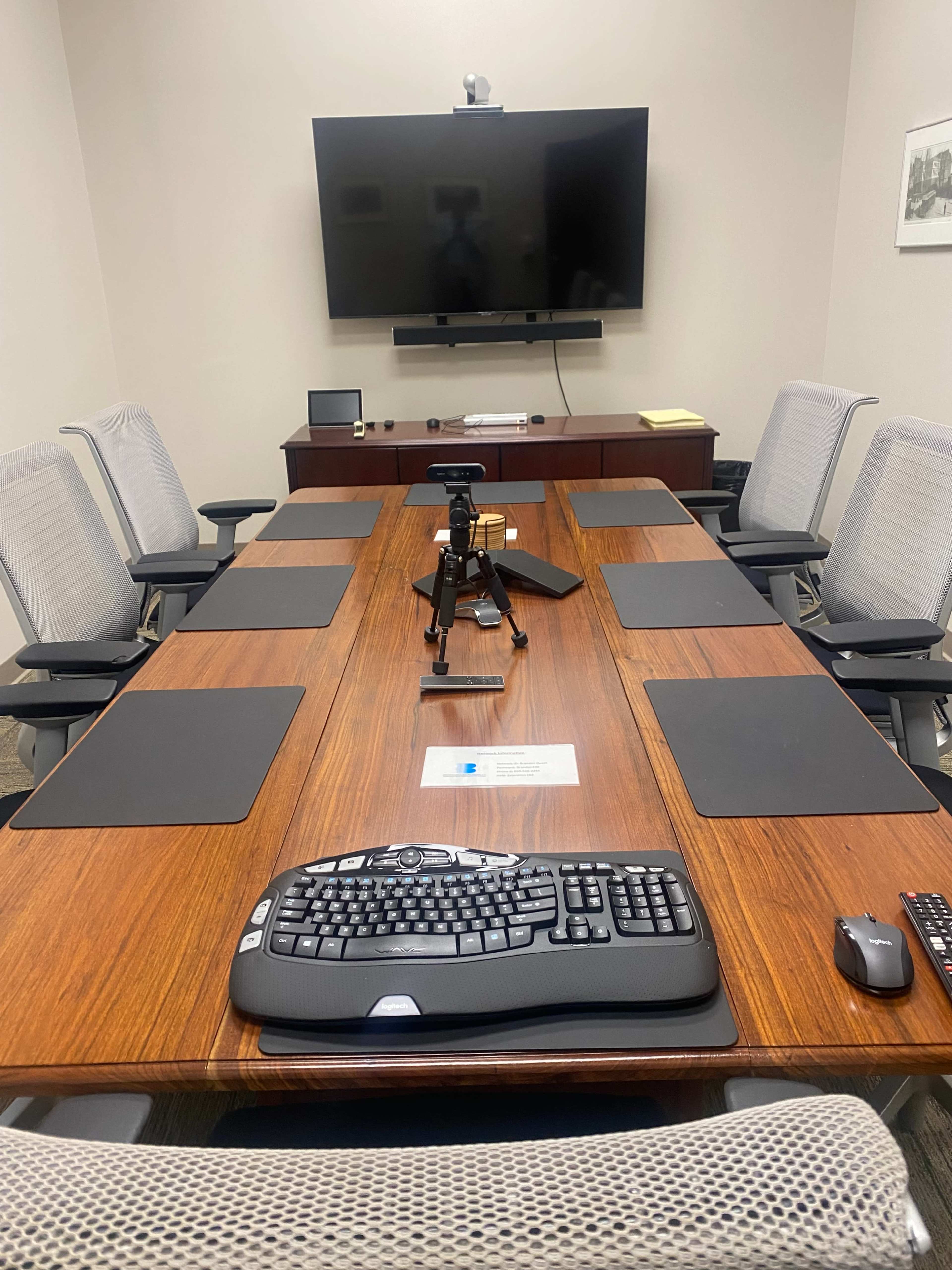 A conference room features a large wooden table surrounded by grey office chairs, with a television screen mounted on the wall and a tripod placed in the center.