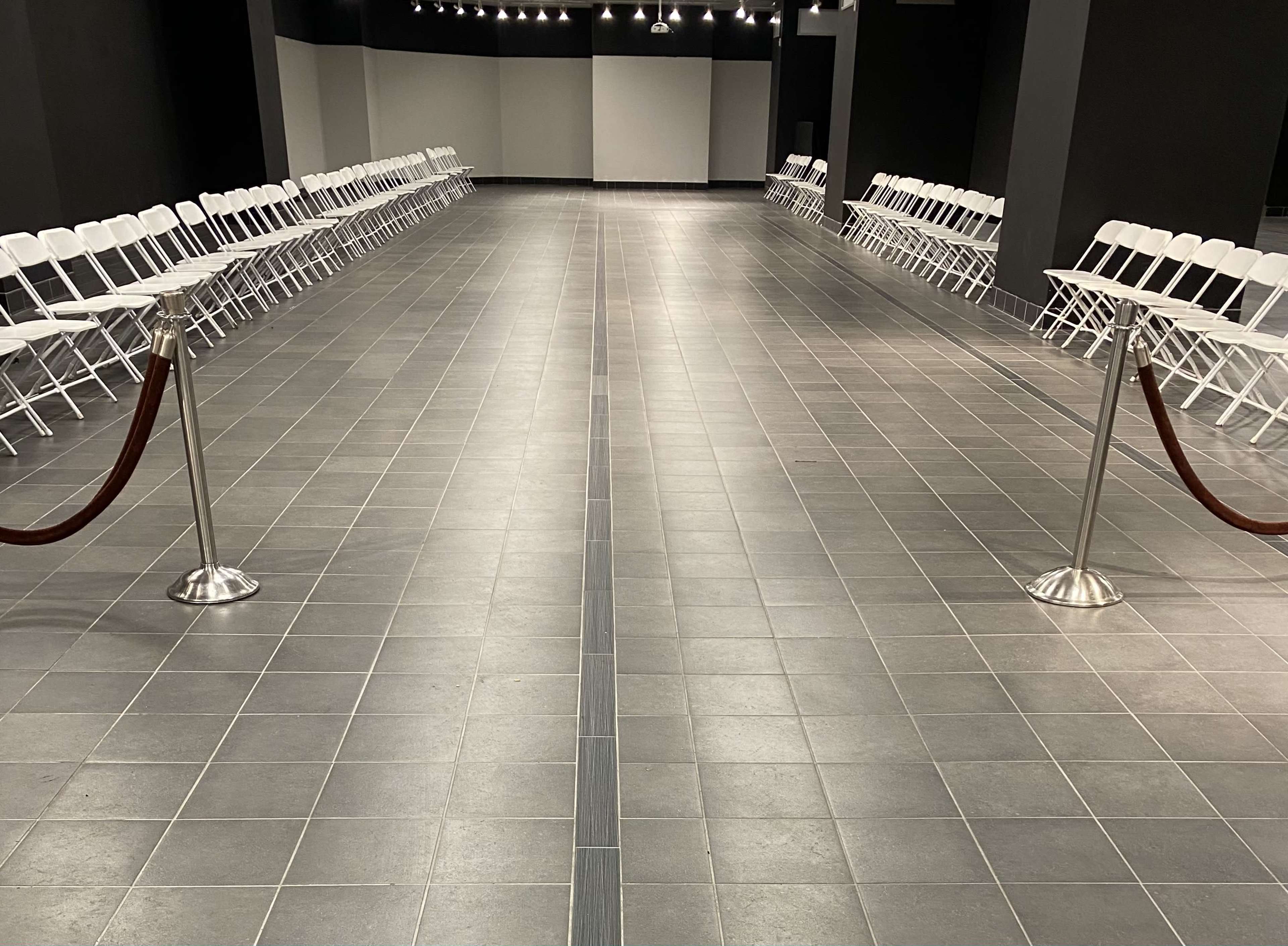 A spacious hall with two rows of white folding chairs arranged on either side of a central walkway, separated by a red rope barrier.