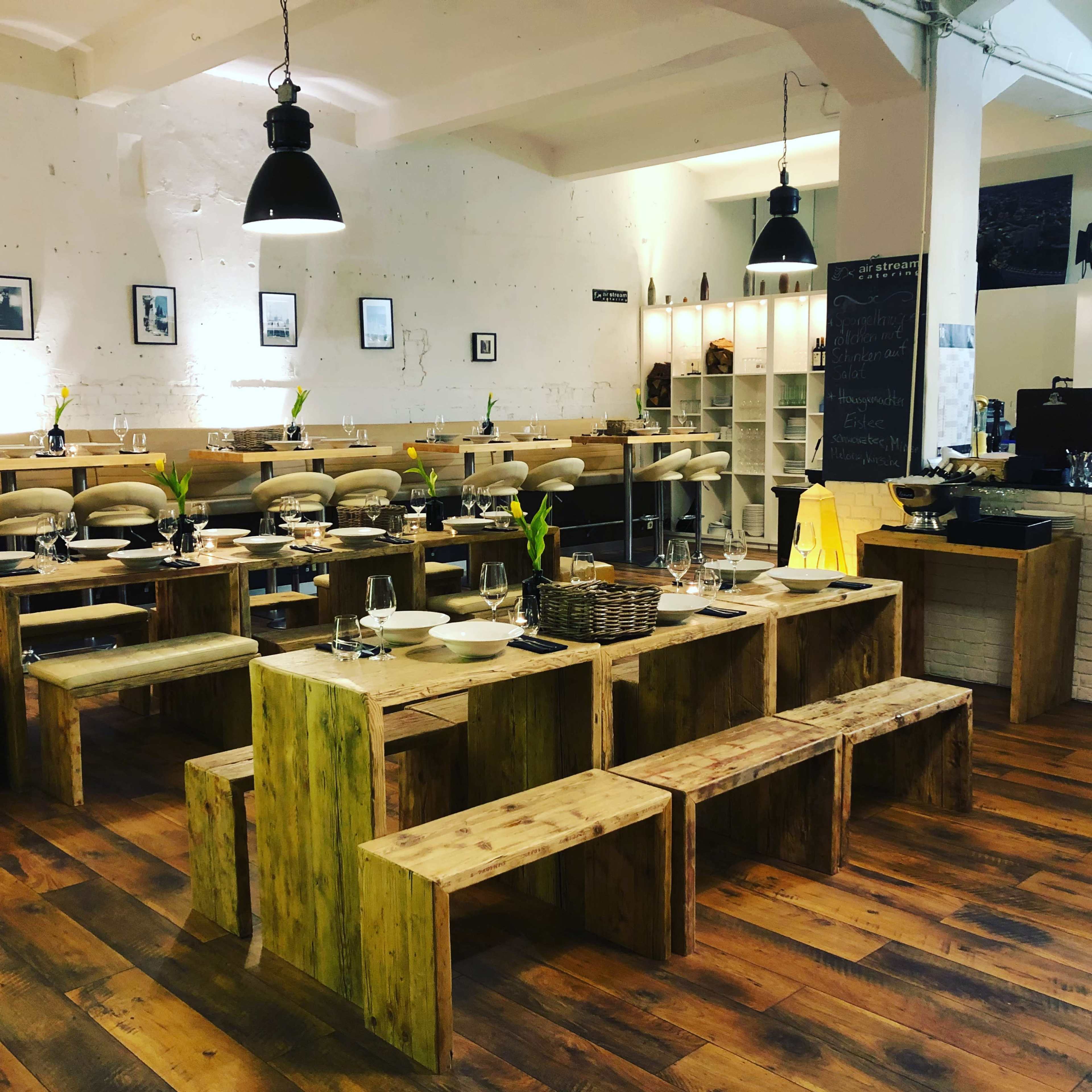 A modern restaurant interior featuring wooden tables and benches, set with dinnerware and flowers, under industrial-style lighting.