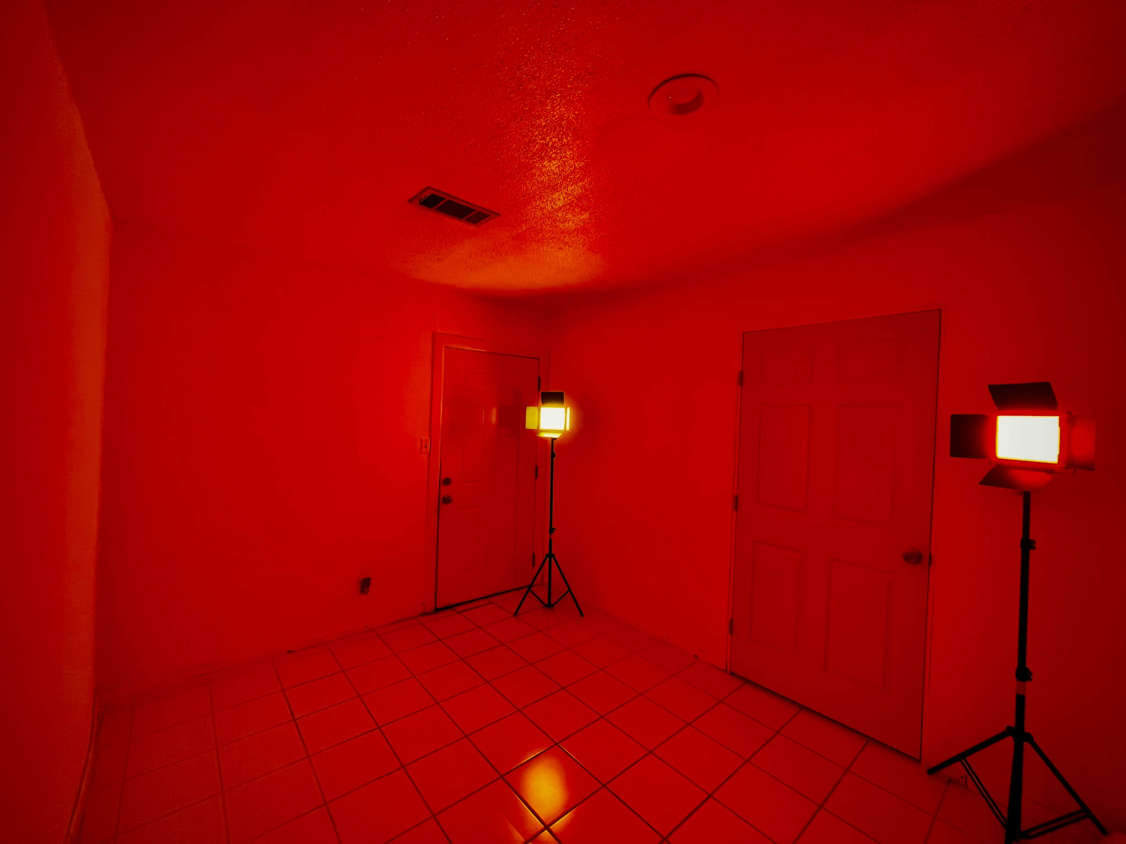 The image shows a small room illuminated by red lights, featuring a door on one wall and a second door on an adjacent wall.