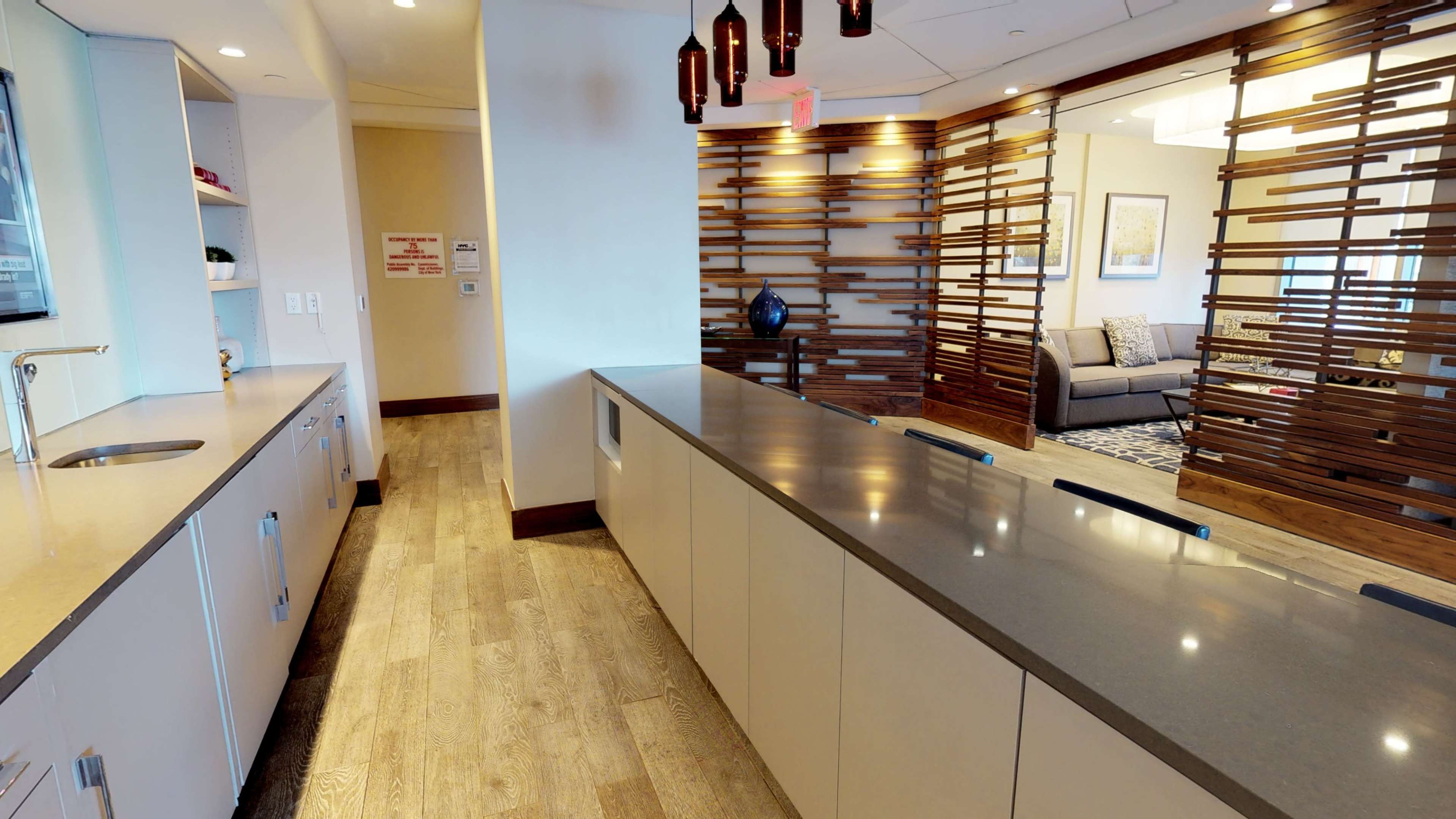 A modern kitchen area with sleek cabinetry and a long countertop adjacent to a lounge area separated by a wooden slatted partition.