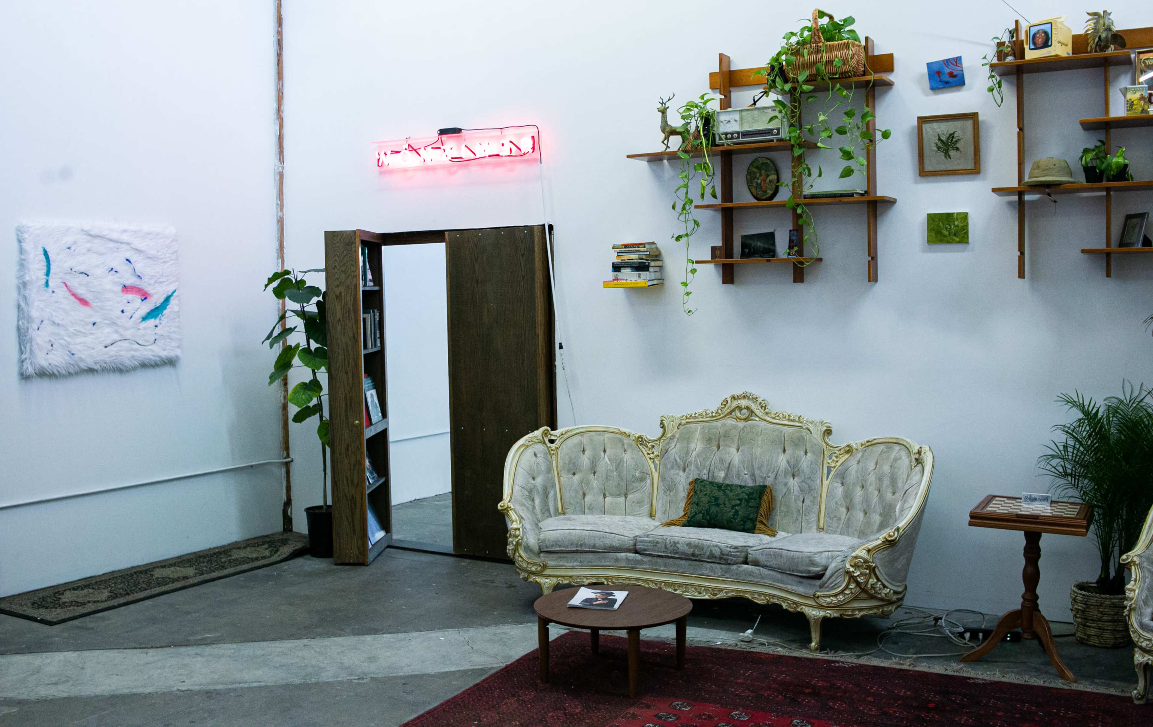 A bright, minimalist room features a vintage couch, wooden tables, shelves with plants and decor, and an open door leading to a bookcase.