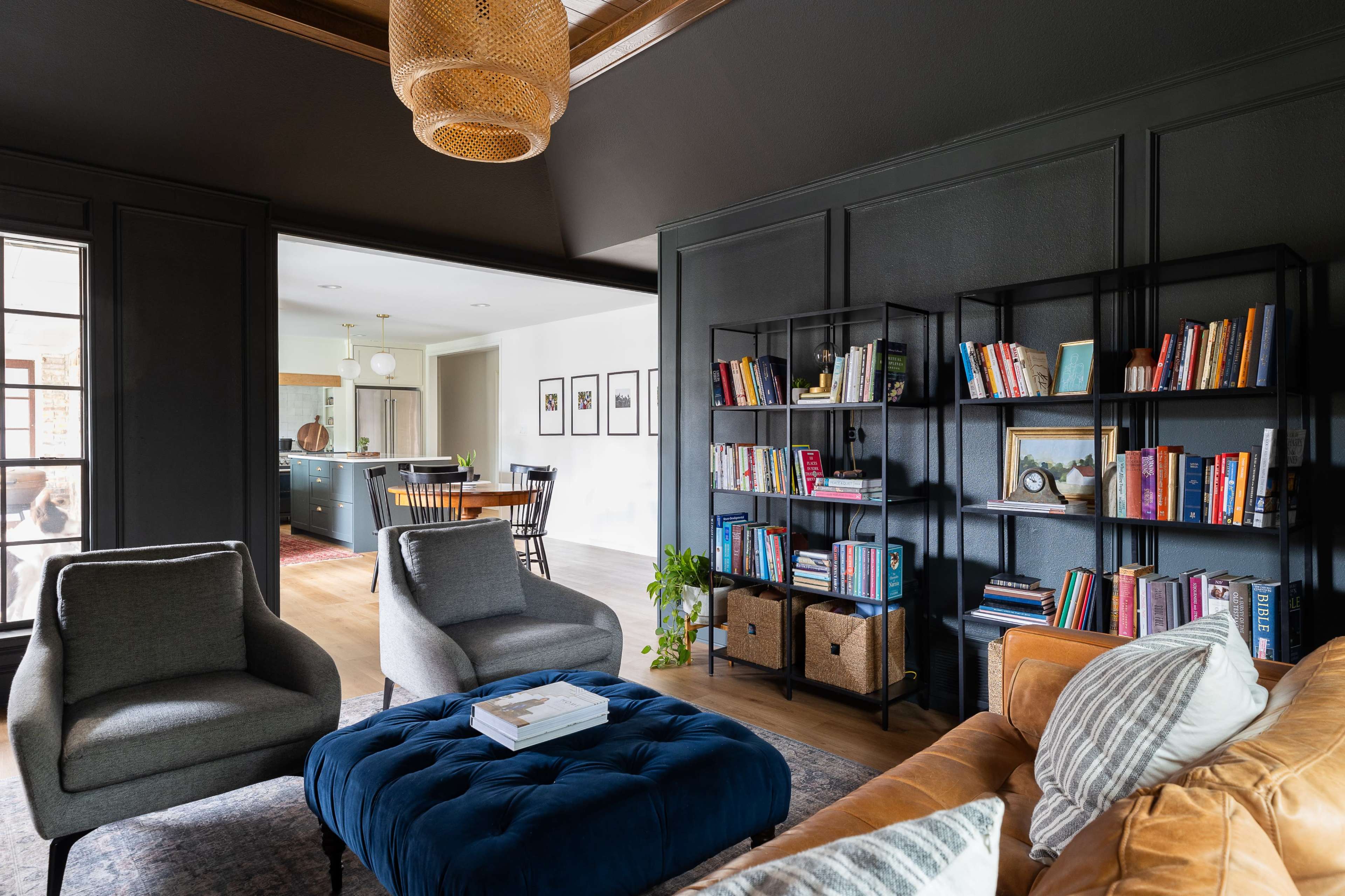 A modern living room features a black accent wall, two gray armchairs, a blue ottoman, a brown sofa, and a bookshelf filled with books, leading into a kitchen area in the background.