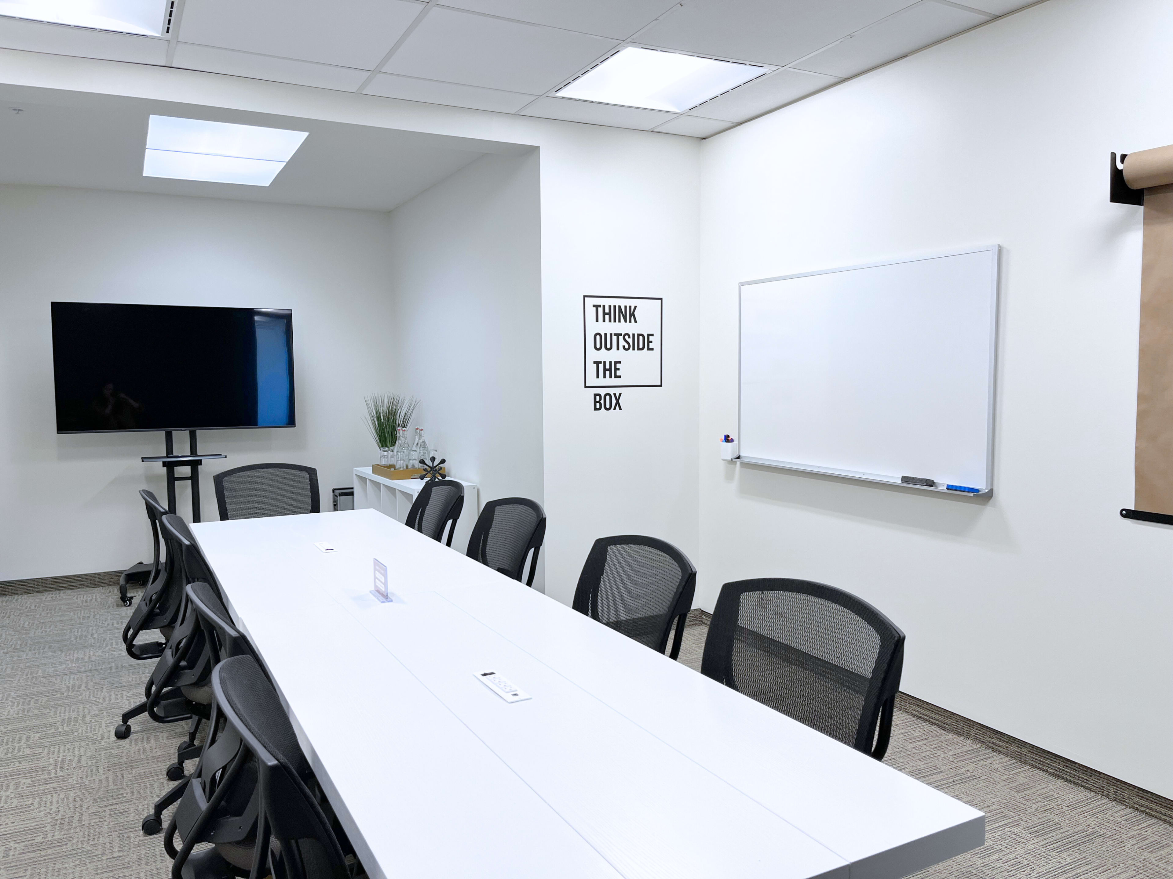 A bright conference room features a large white table, a TV screen, a whiteboard, and a motivational poster that says "THINK OUTSIDE THE BOX."