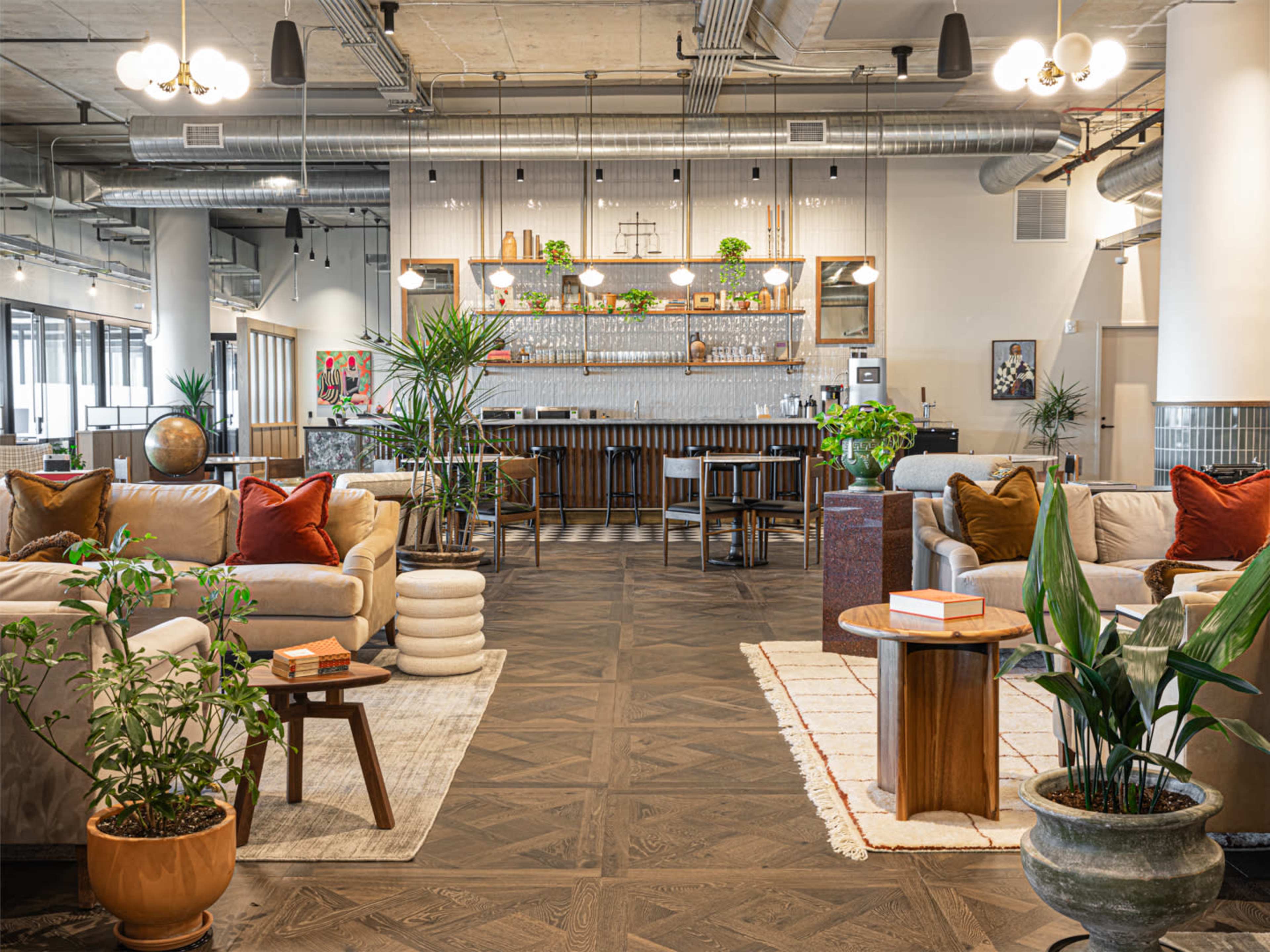 A contemporary lounge area features comfortable sofas, a wooden bar with shelves of decor, and various plants grouped around tables.