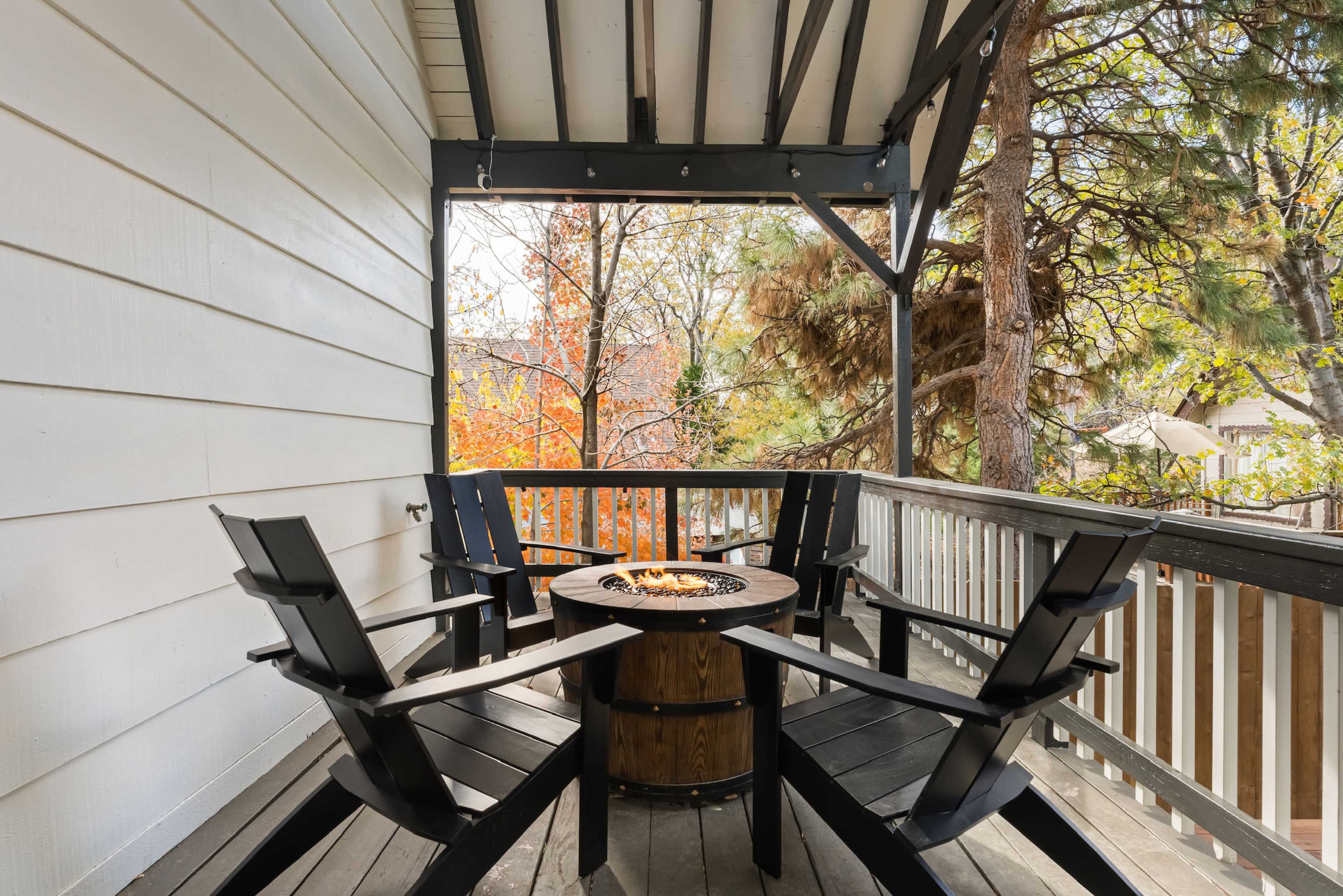 A wooden deck featuring four black adirondack chairs arranged around a circular fire pit.