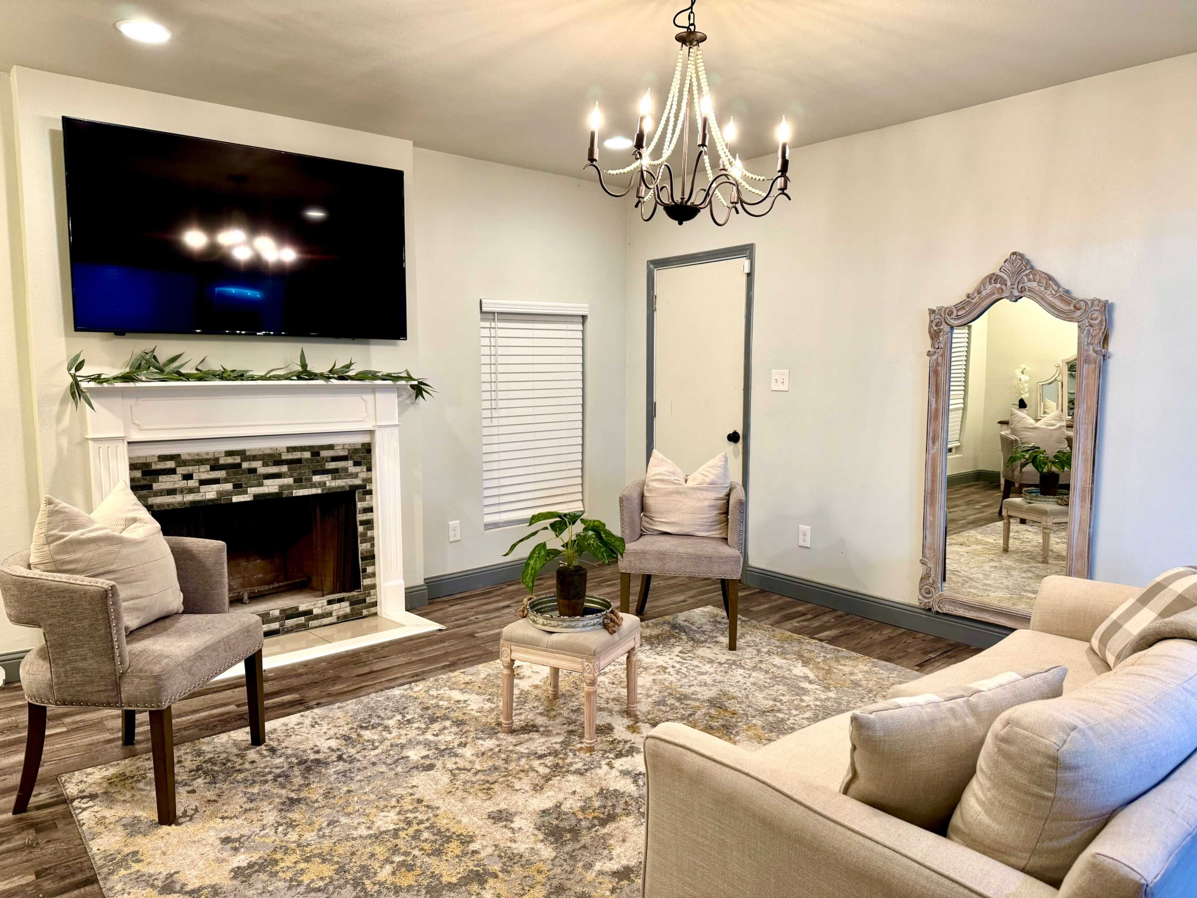 The image shows a cozy living room with a fireplace, chairs, a sofa, a large wall-mounted TV, and a decorative mirror.