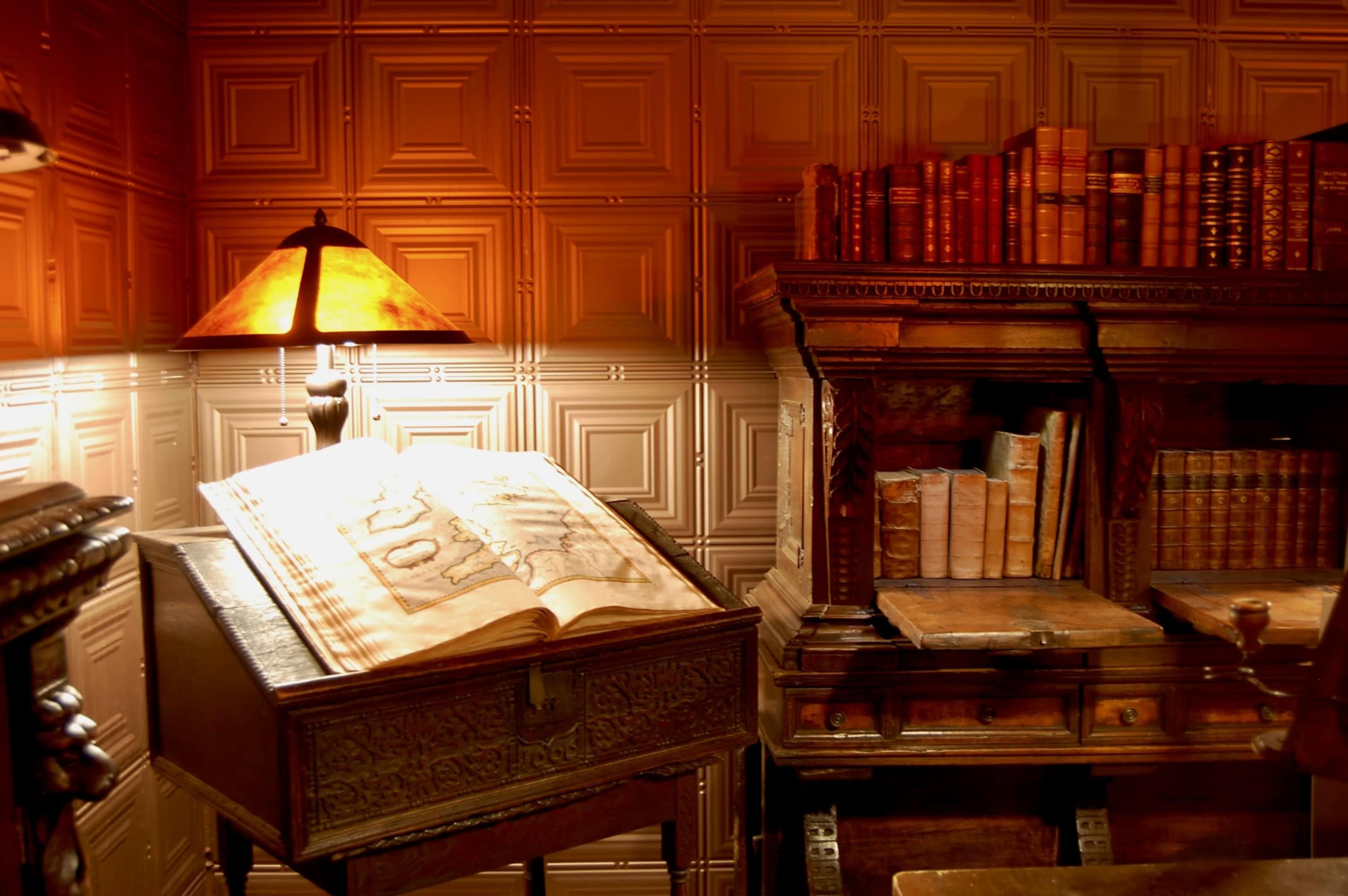 A vintage reading room featuring a large open book on a decorative table, a lamp casting warm light, and several stacks of books on a nearby shelf.