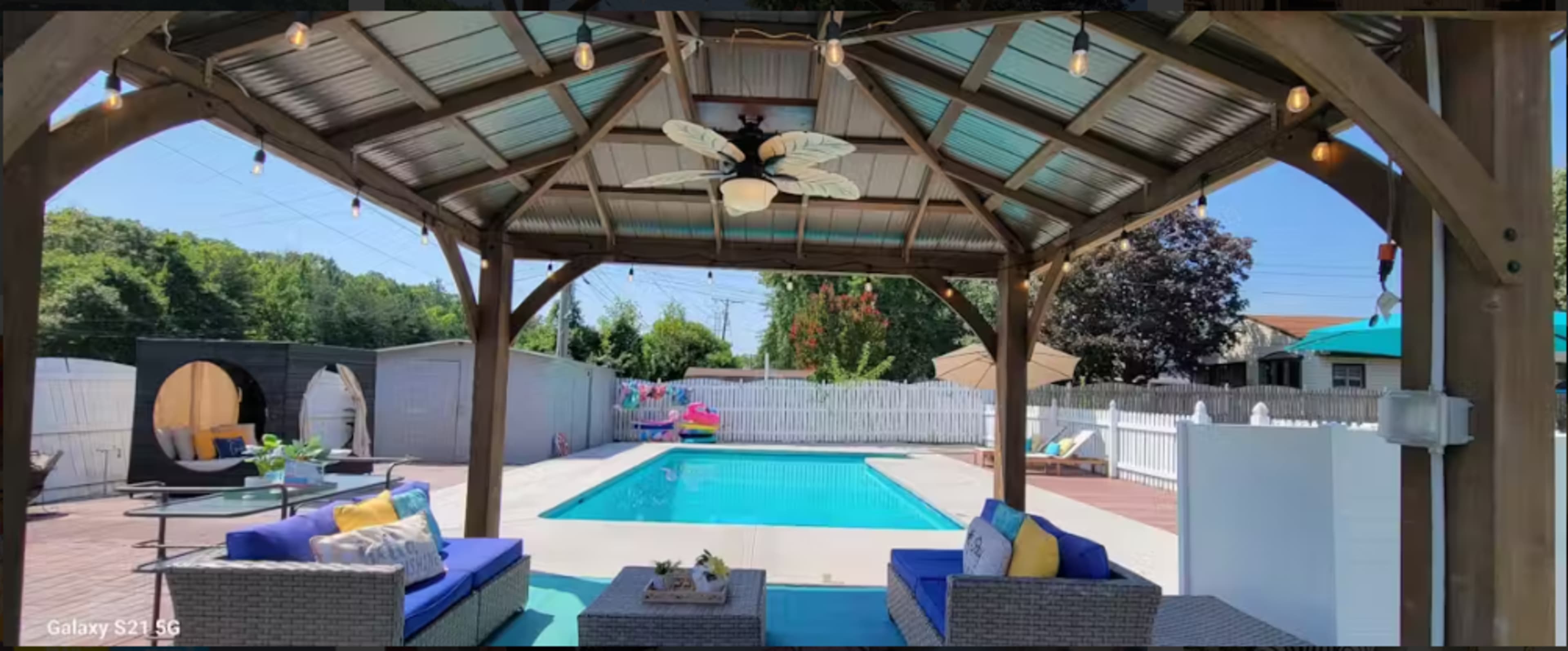 The image shows a shaded outdoor seating area with blue couches, facing a swimming pool surrounded by a patio and a fence.