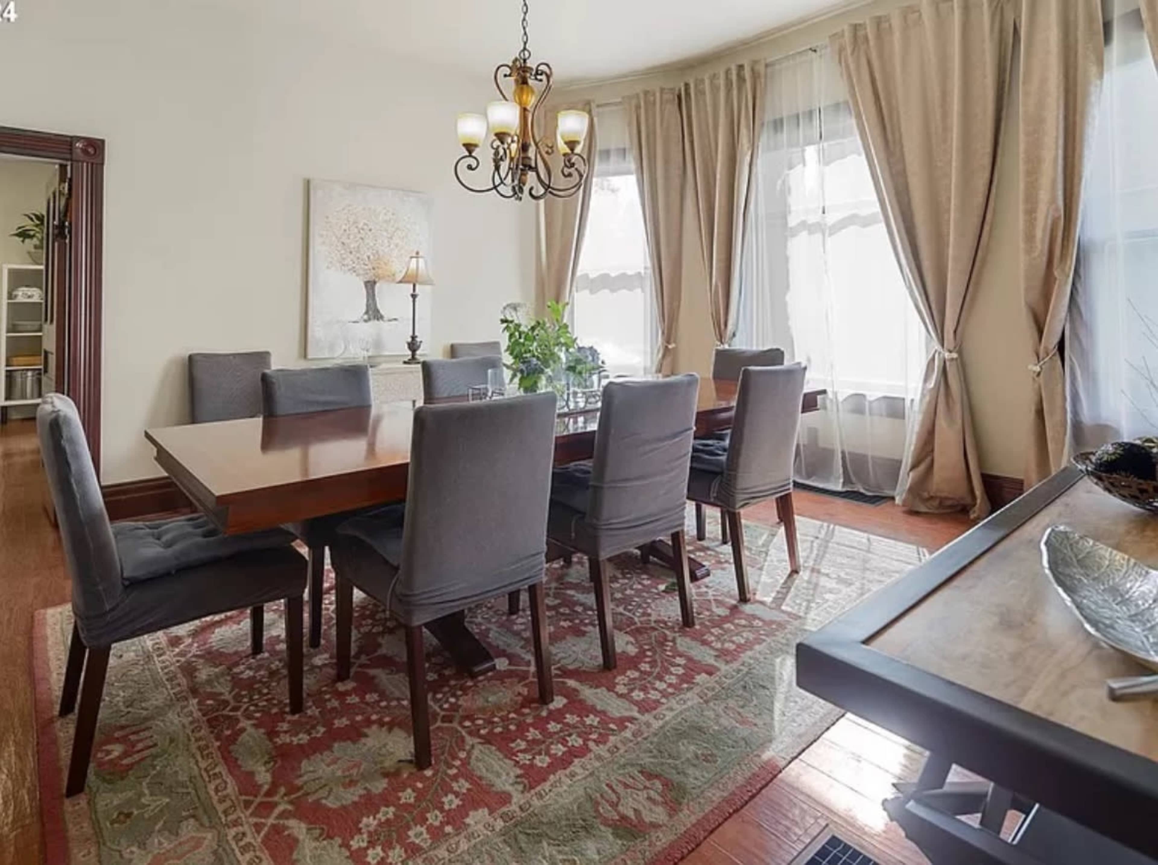 A dining room features a large wooden table surrounded by eight upholstered chairs, with large windows draped in curtains and a decorative chandelier overhead.