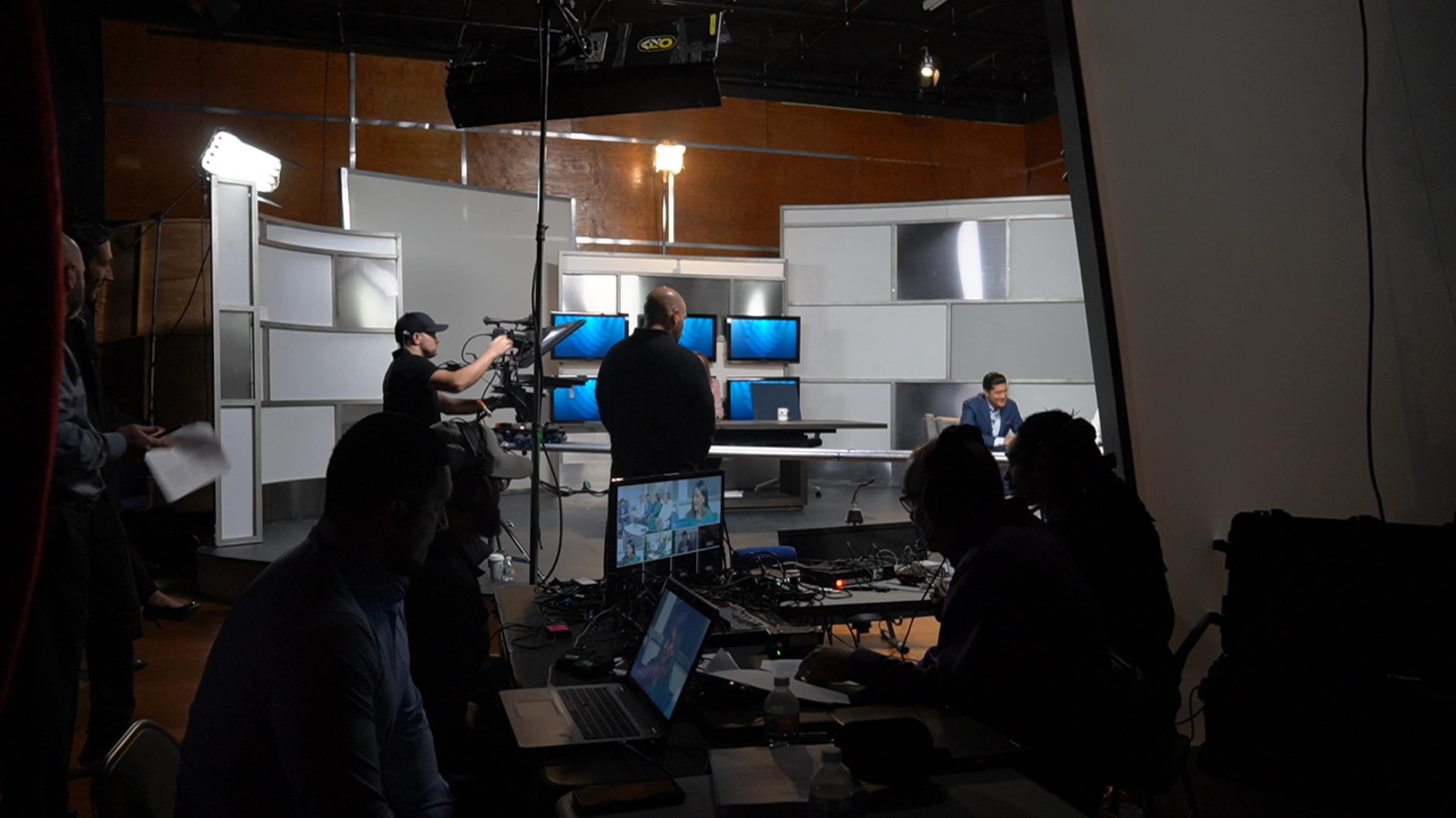 The image shows a television studio setup with multiple screens, cameras, and crew members preparing for a broadcast.