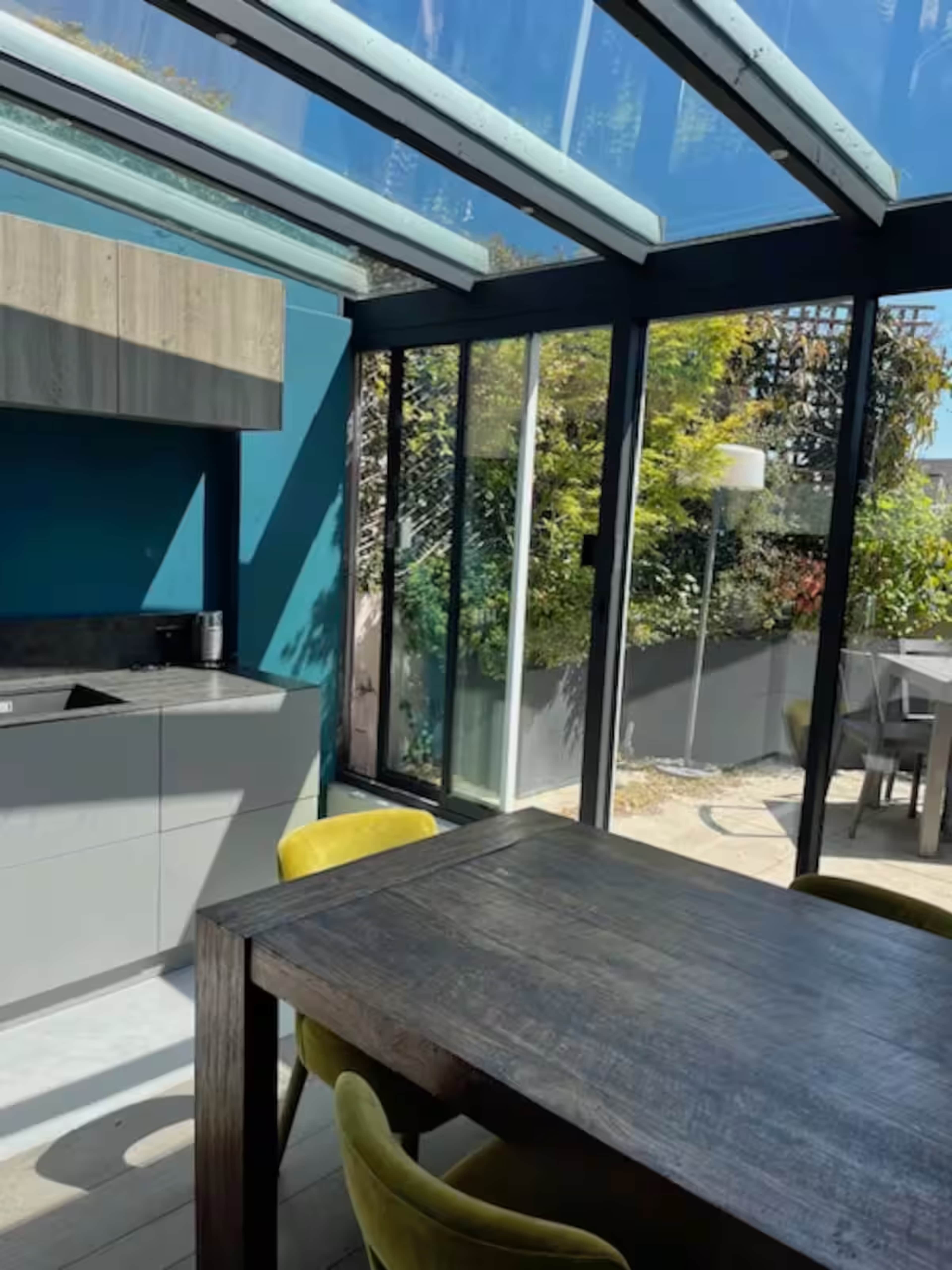 The image shows a modern kitchen and dining area with large glass windows and a wooden table surrounded by yellow chairs.