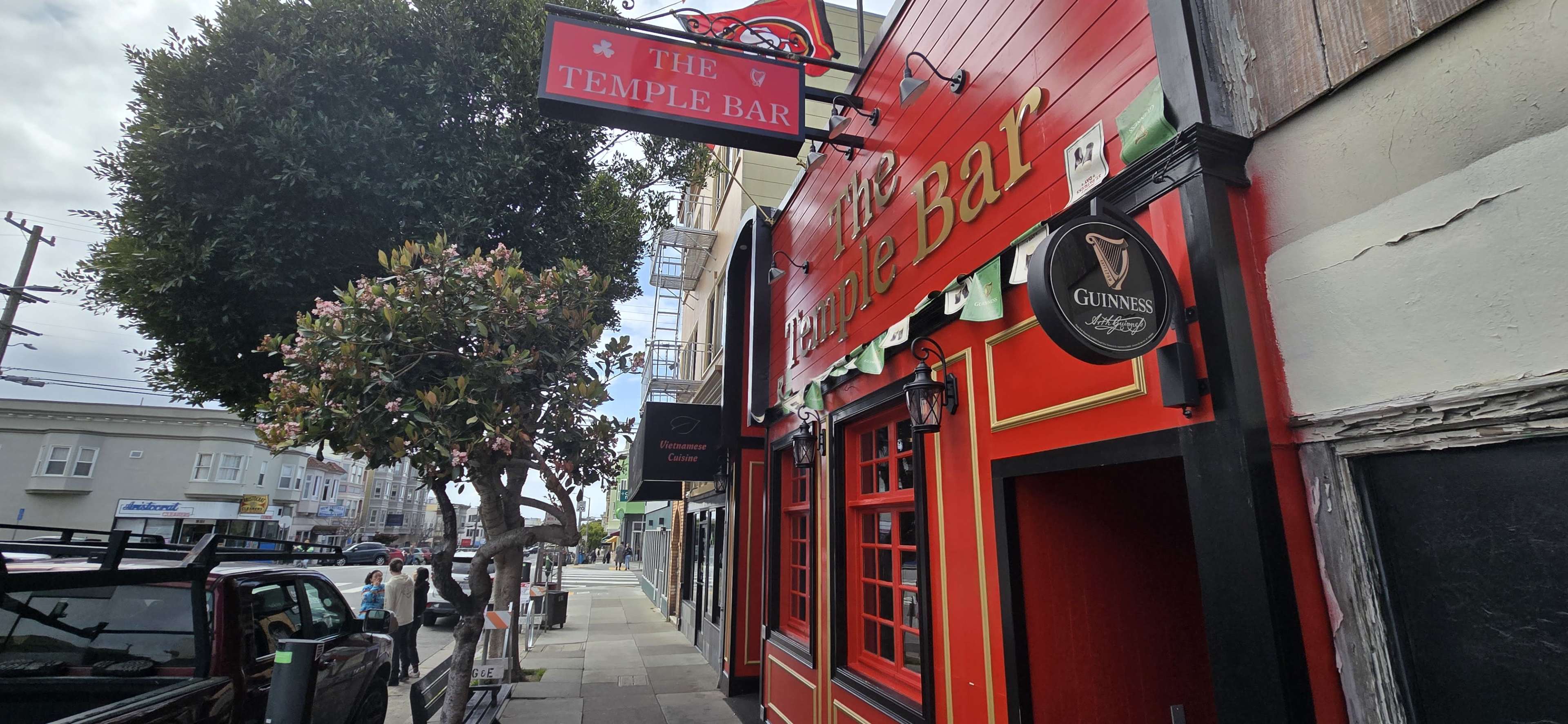 The image shows the exterior of a bar named "The Temple Bar," featuring a bright red facade and a sign displaying its name, alongside a tree and parked cars on the street.