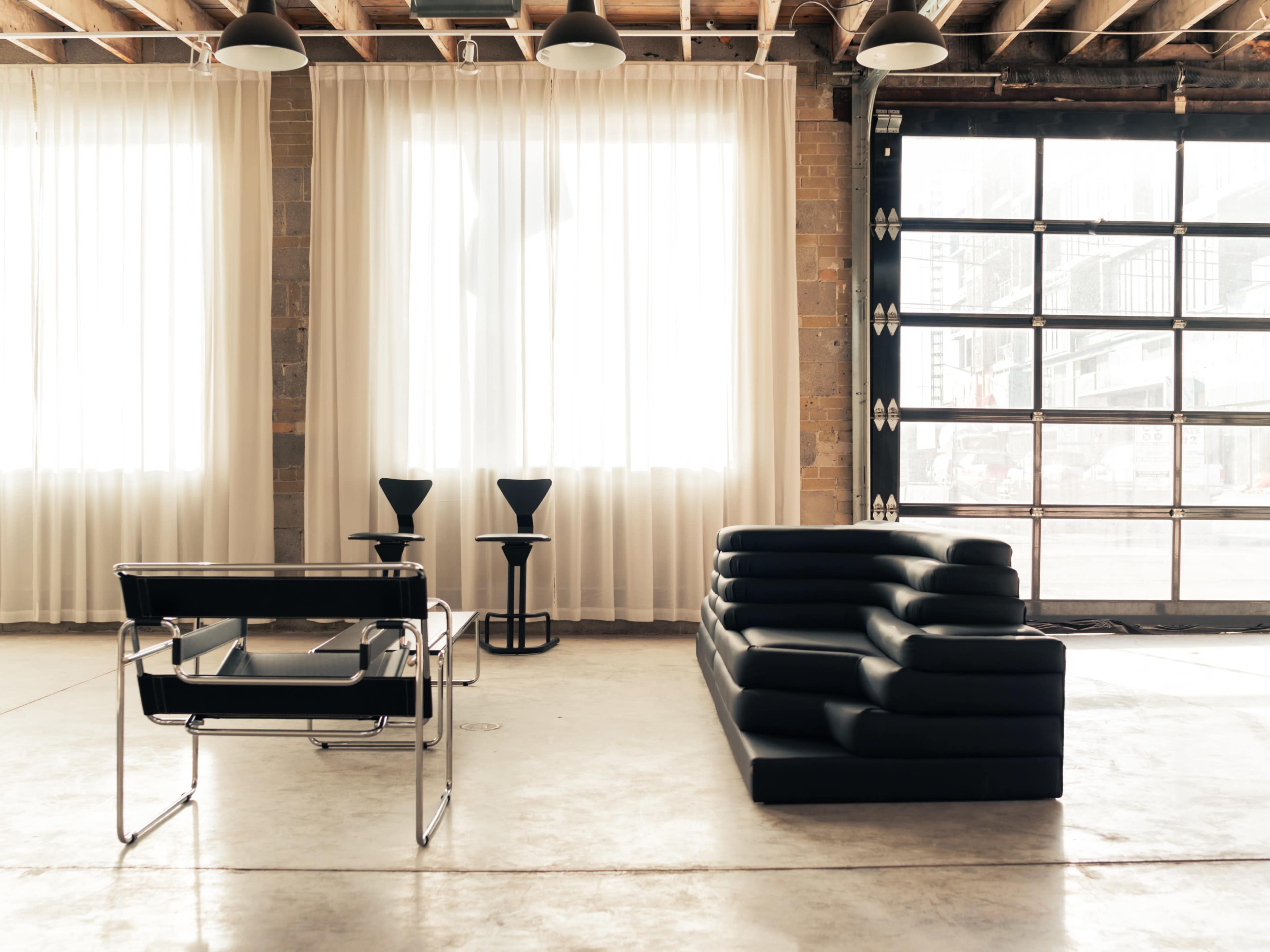 The image shows a modern interior space featuring a black sectional sofa, two small black tables, and large windows with sheer curtains.